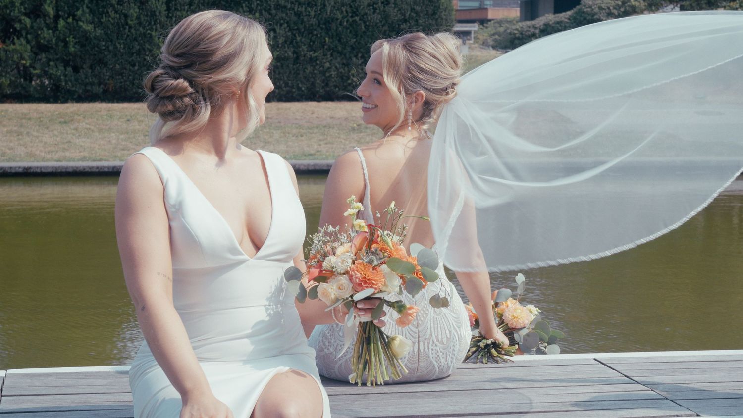 Two brides in white wedding dresses sit on a dock holding bouquets, one veil blowing in the wind.