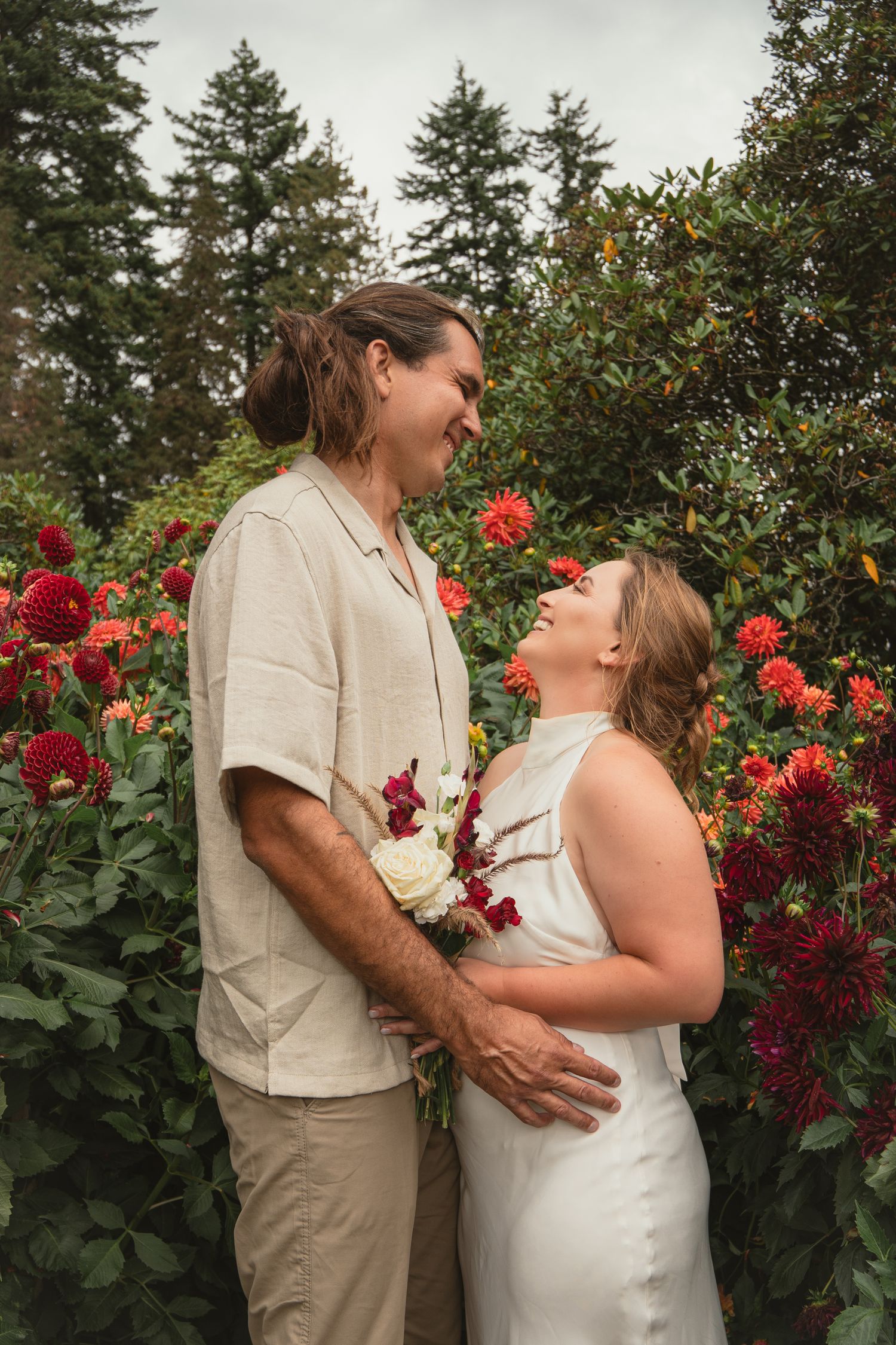 Couple gazing at each other lovingly in a garden surrounded by vibrant red dahlia flowers.