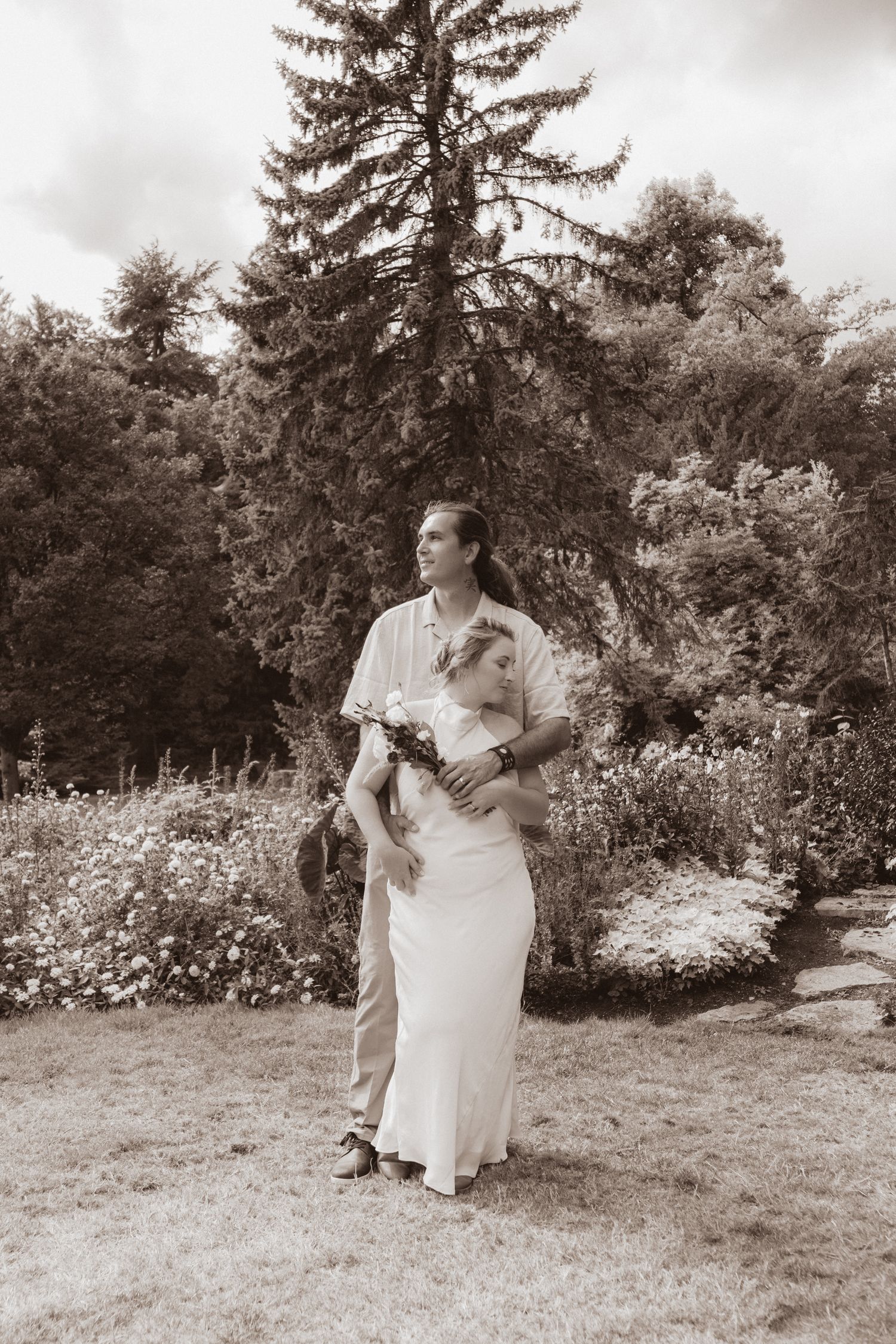 Sepia-toned portrait of bride and groom standing together in a garden, bride holding a bouquet.