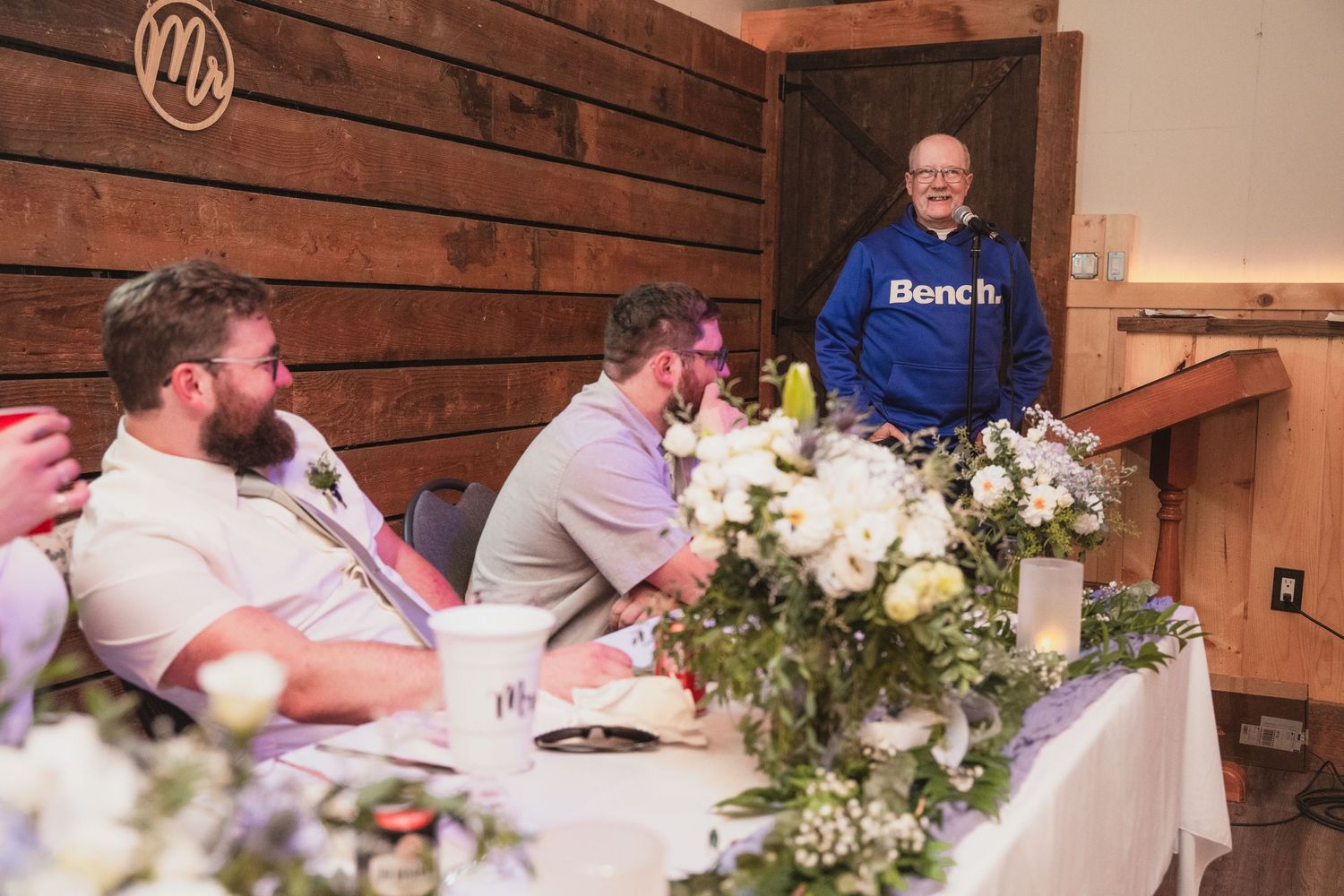 Man in blue Bench hoodie giving a speech at a wedding reception in a rustic barn venue.