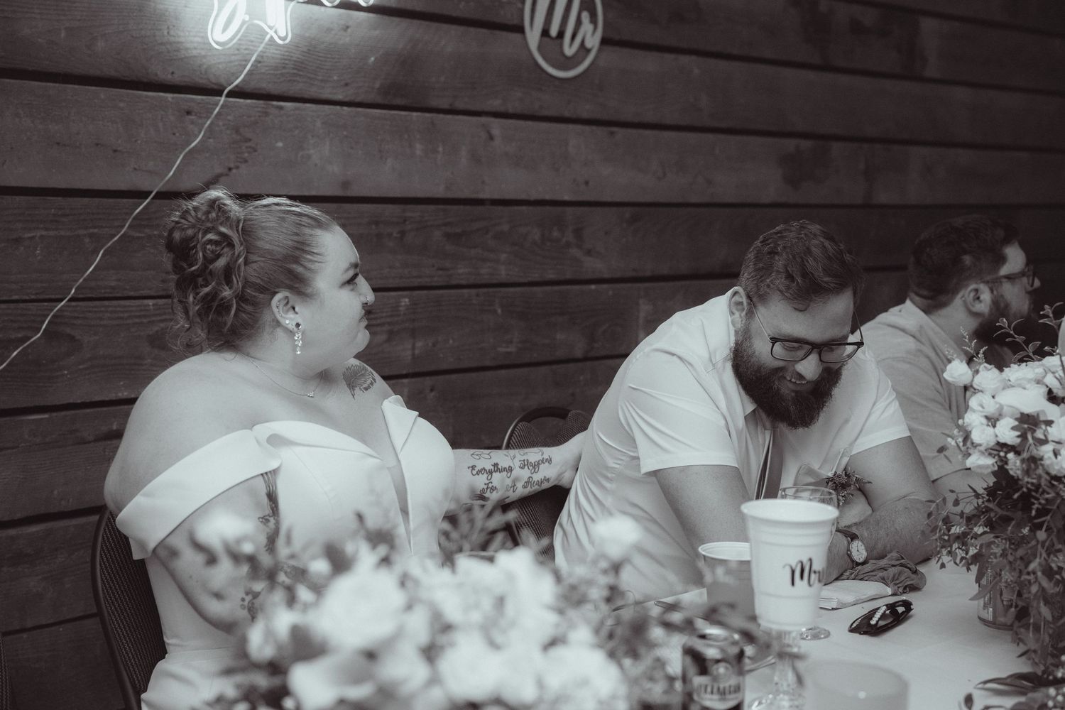 Black and white wedding reception photo of bride and groom seated at table with floral centerpieces.