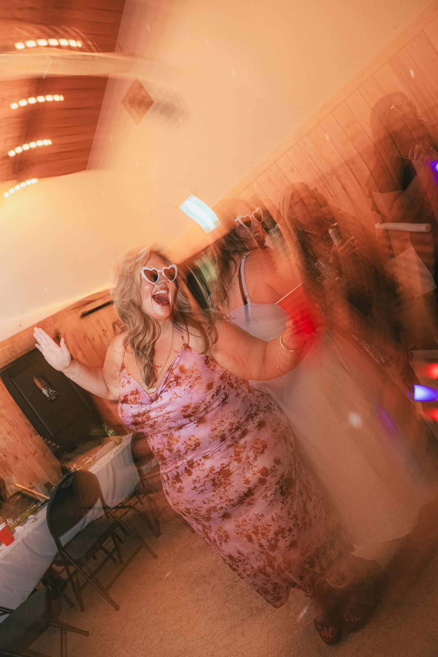 Woman in floral dress and heart sunglasses dancing joyfully at an indoor party with warm lighting.