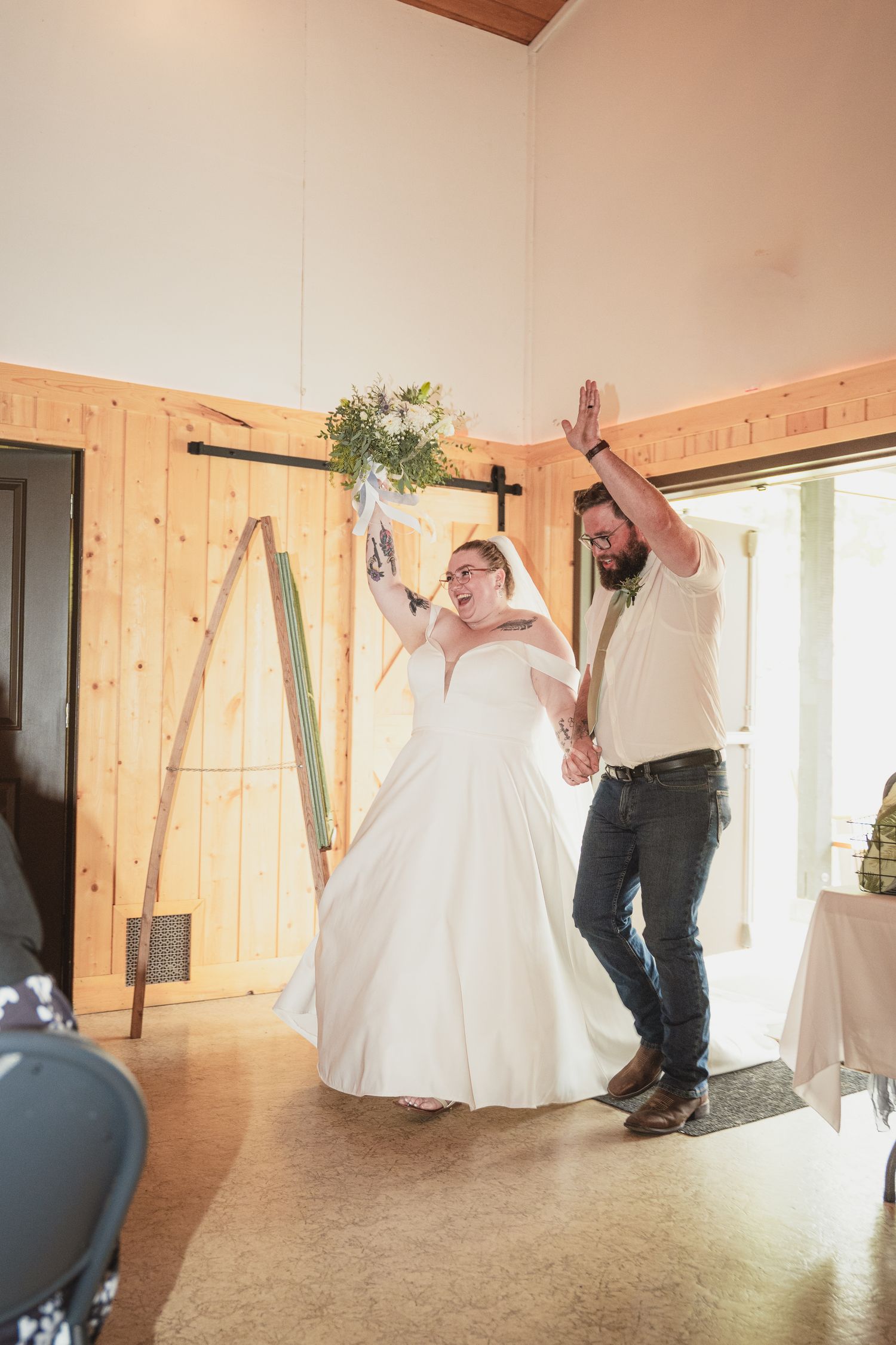Joyful newlyweds make grand entrance into rustic barn wedding venue, bride in white gown raising bouquet, groom in jeans celebrating.