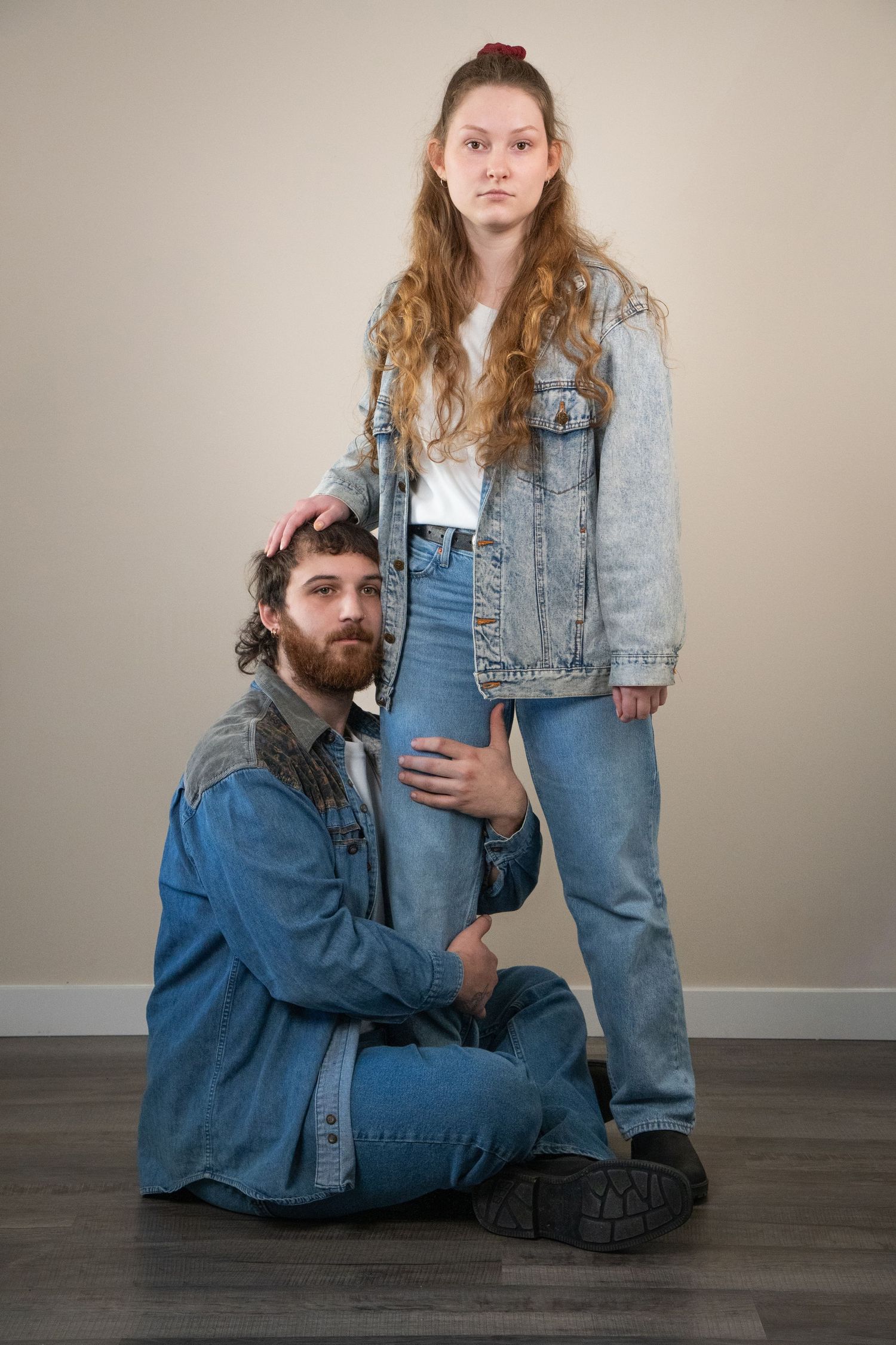 A bearded man sits on the floor holding a young womans leg as she stands beside him in denim.