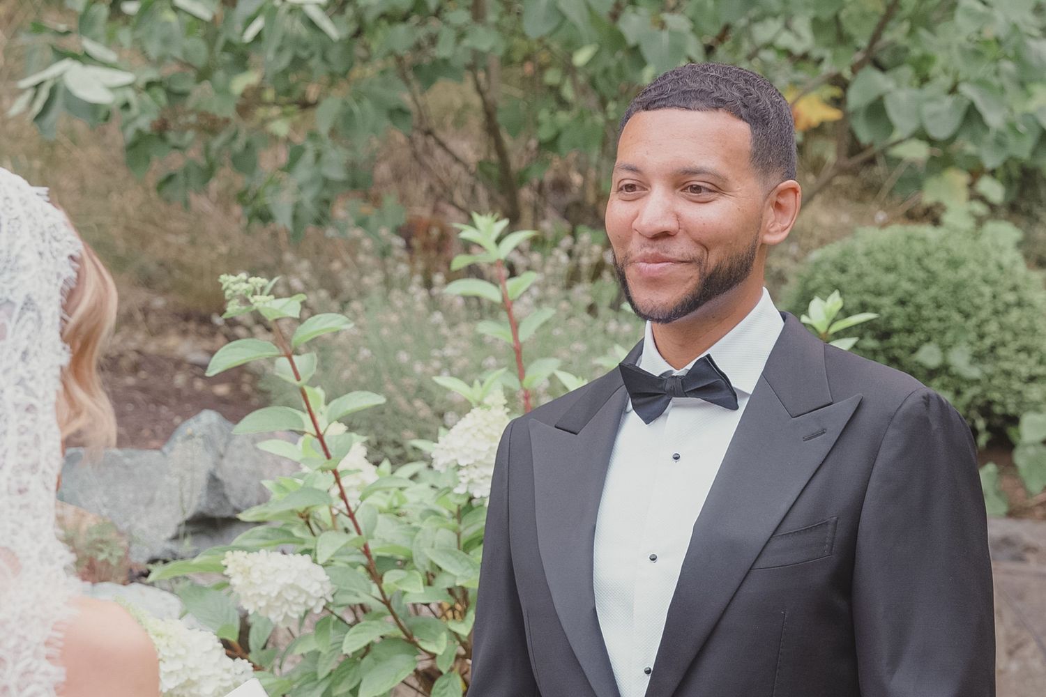 Smiling groom in a black tuxedo watching his bride approach during an outdoor garden wedding ceremony.