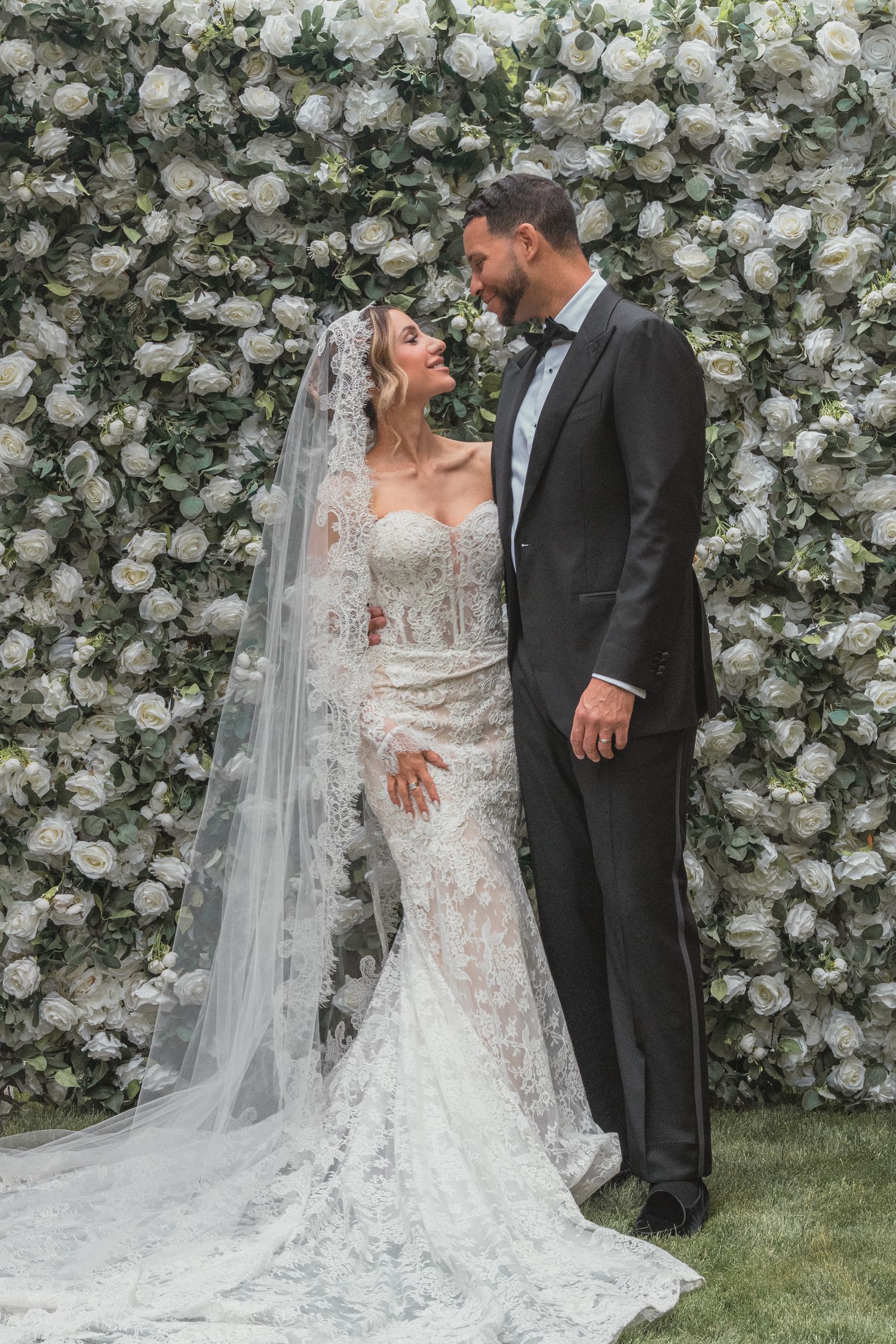 Bride and groom gazing at each other in front of a white rose flower wall, bride wearing lace gown and veil.