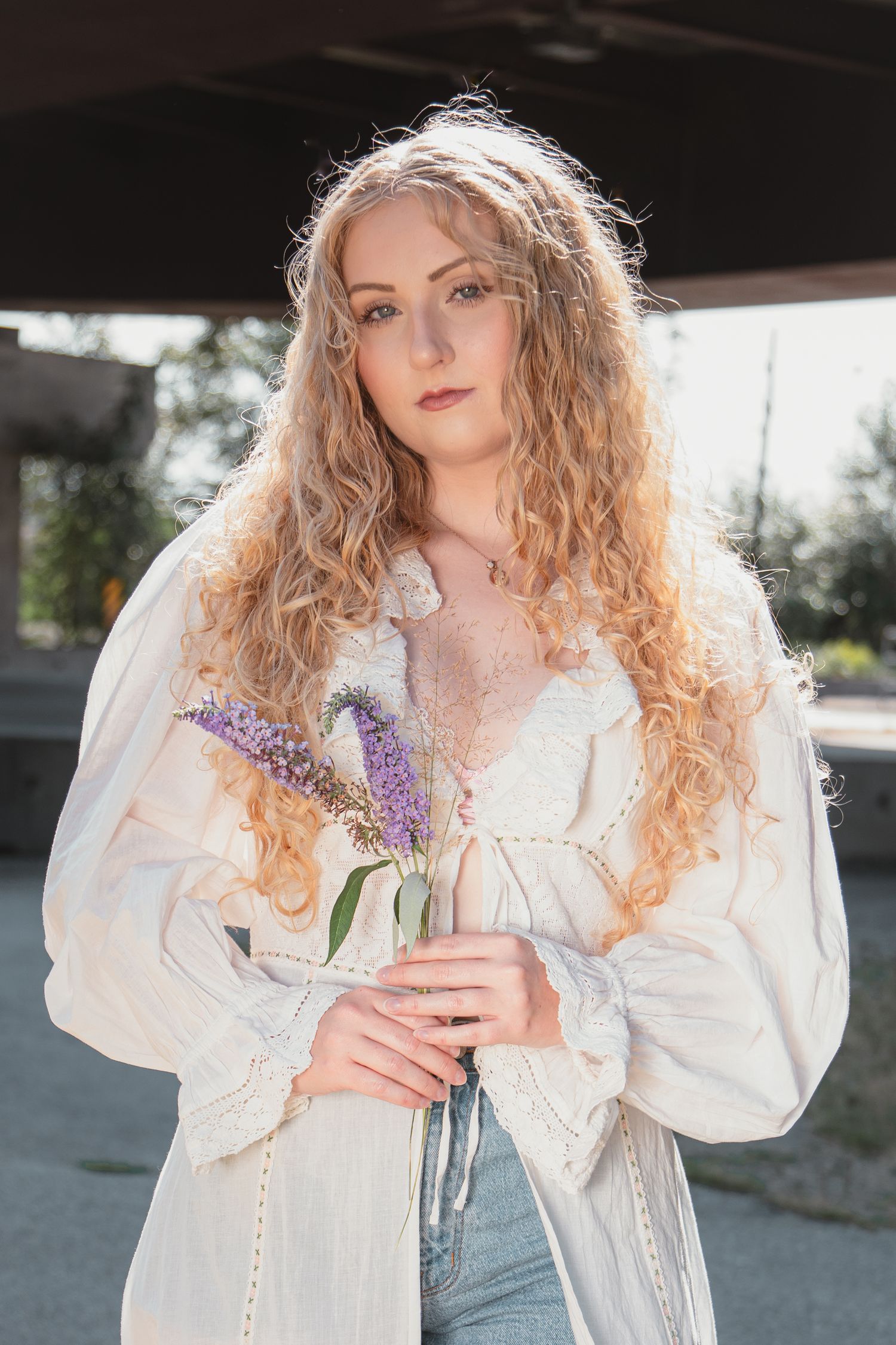 Young woman with curly blonde hair holding purple lavender flowers, wearing a white boho blouse and jeans outdoors.