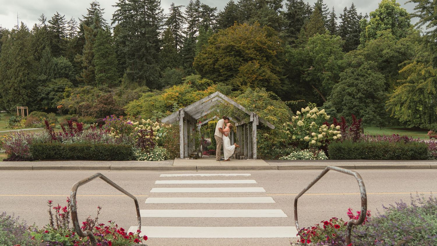 Couple sharing a kiss under a garden arbor surrounded by lush greenery and colorful flowers at an outdoor wedding venue.