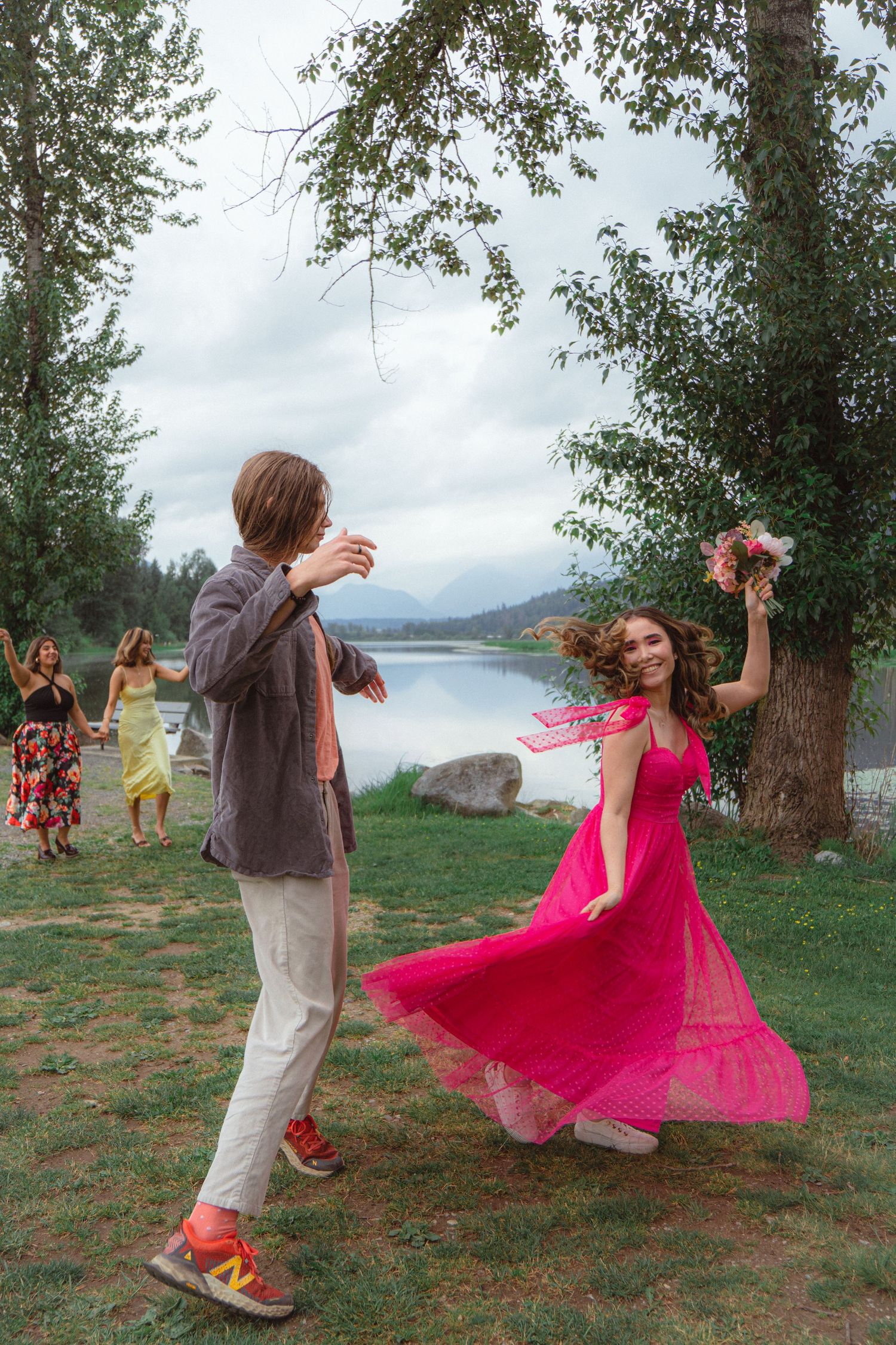 A couple dances joyfully near a lake with guests celebrating in the background.