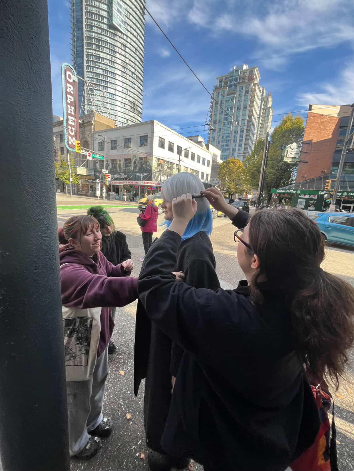 Students adjust a blue wig on a person outdoors on a busy city street on a sunny day.
