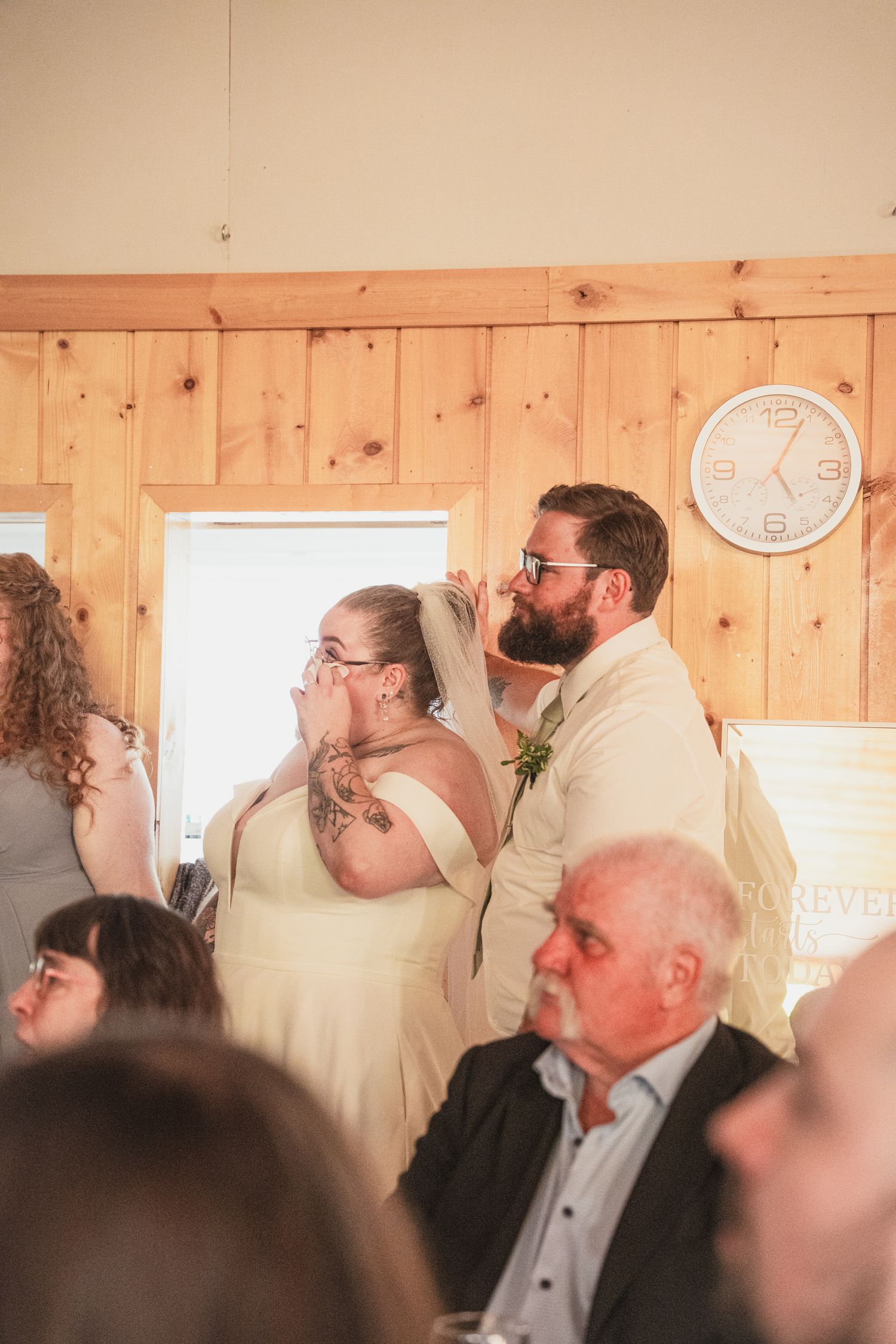 Emotional bride wiping tears while groom comforts her during wedding reception in rustic wood-paneled venue.