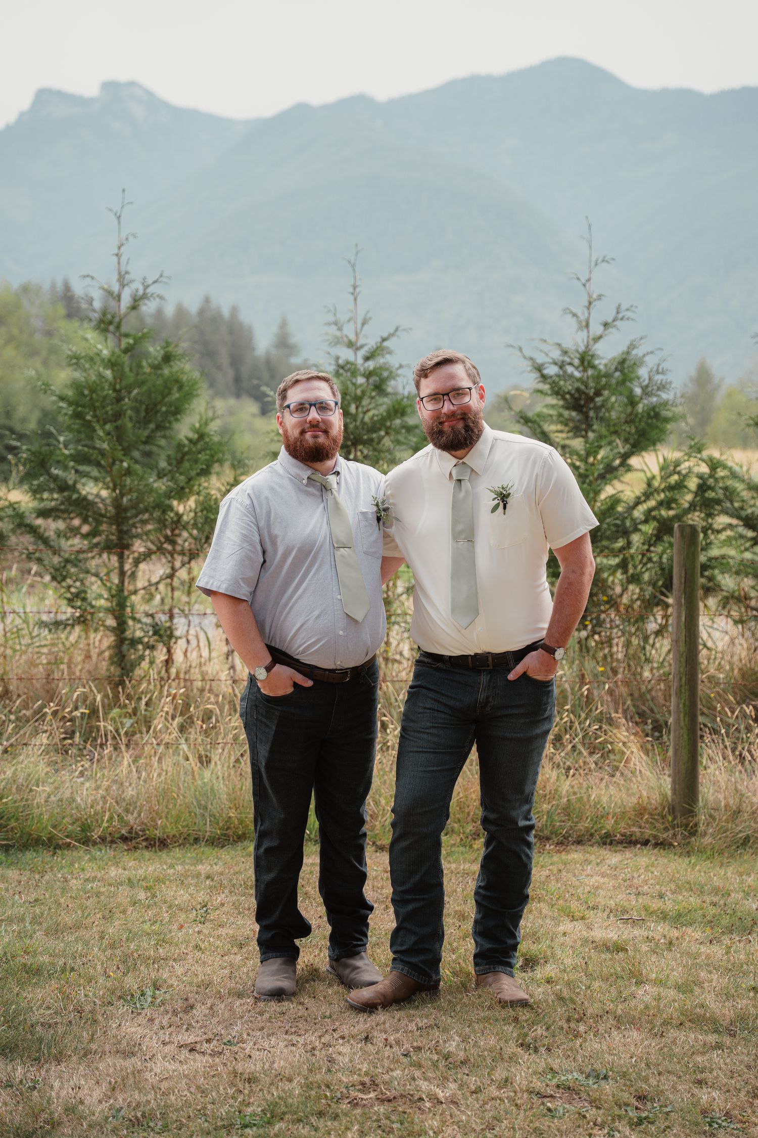Two bearded men in dress shirts and ties pose outdoors with mountain scenery at a wedding.