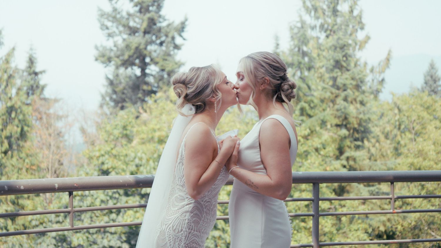 Two brides in white gowns share a kiss on an outdoor balcony surrounded by lush green trees.