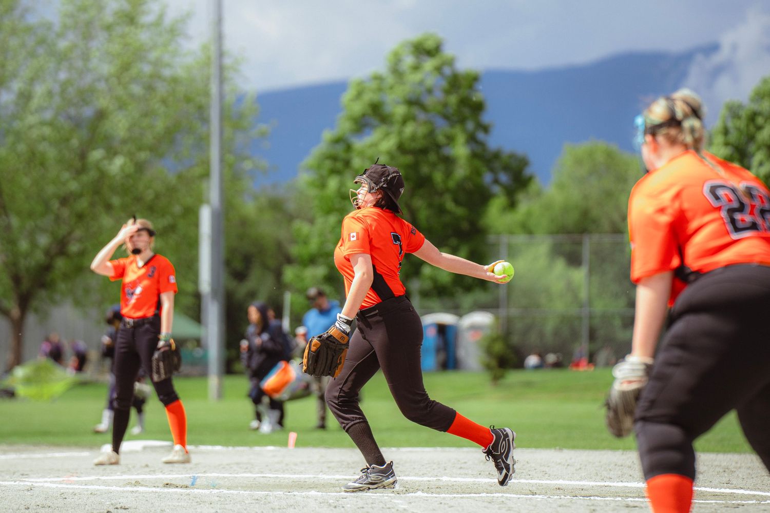 Female softball pitcher in orange uniform delivers a pitch during an outdoor game with teammates nearby.
