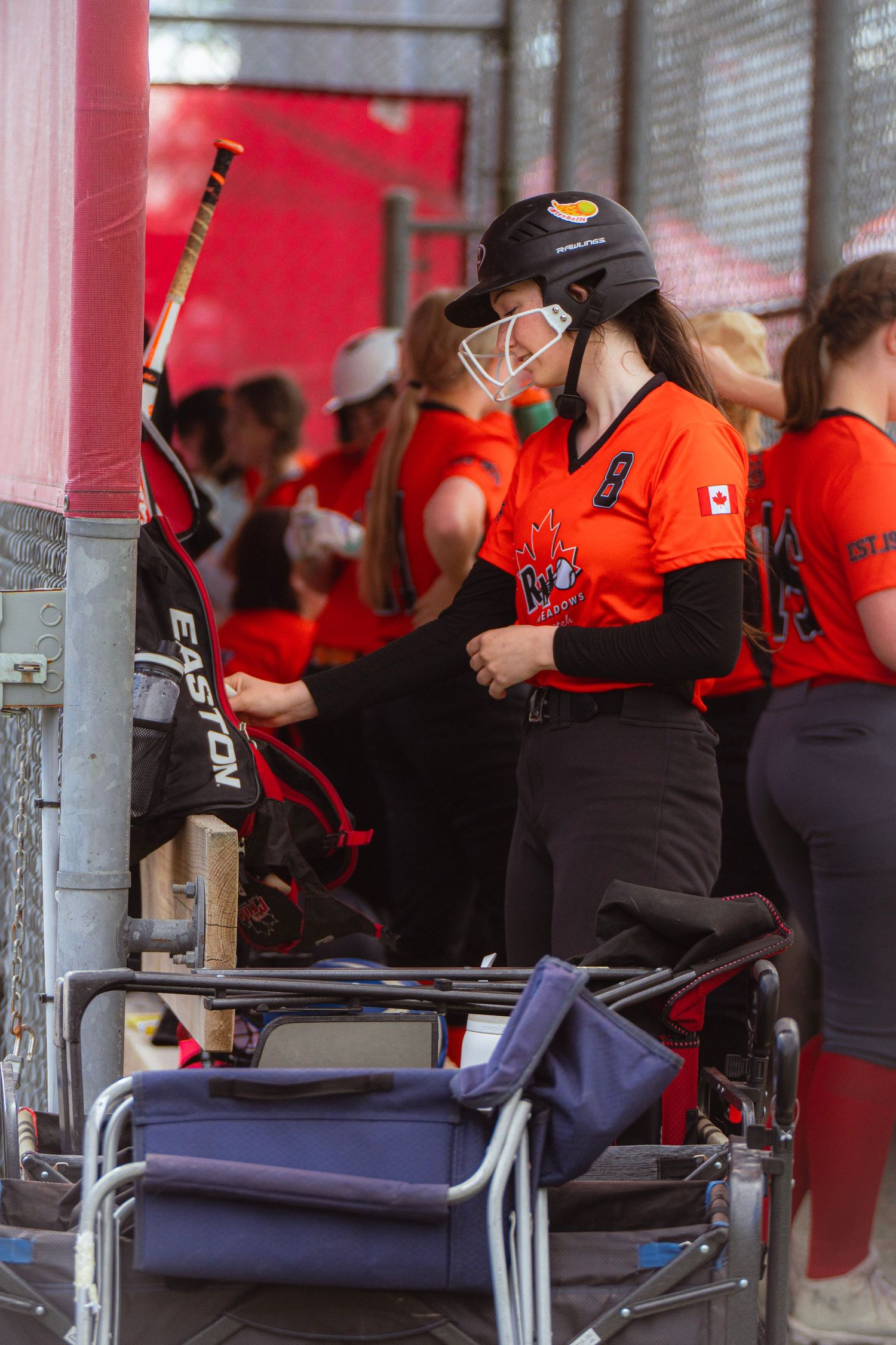 Female softball player in orange uniform preparing equipment in dugout before game.