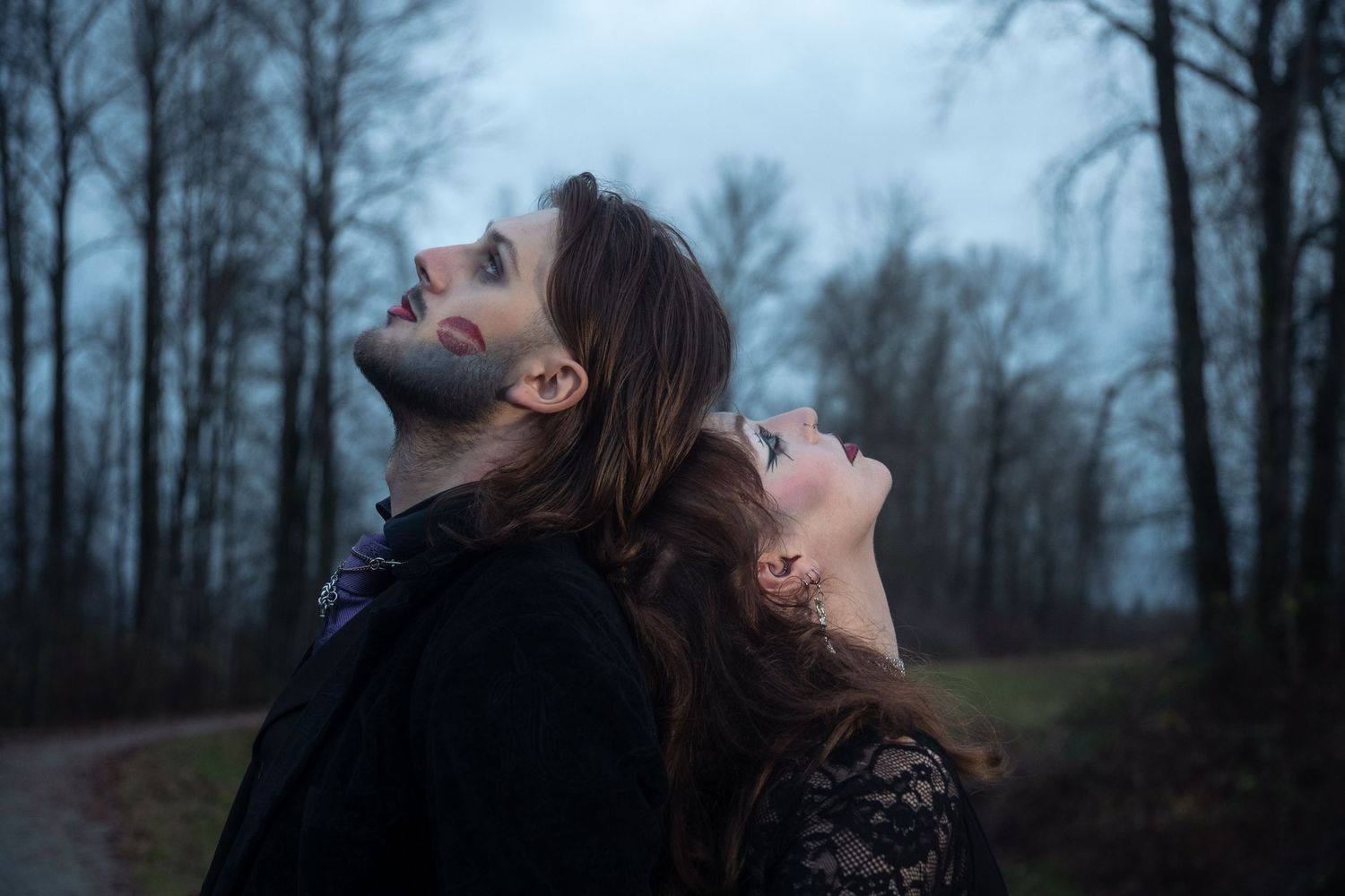 A gothic couple in dark clothing stand back-to-back on a forest path, both gazing upward dramatically.