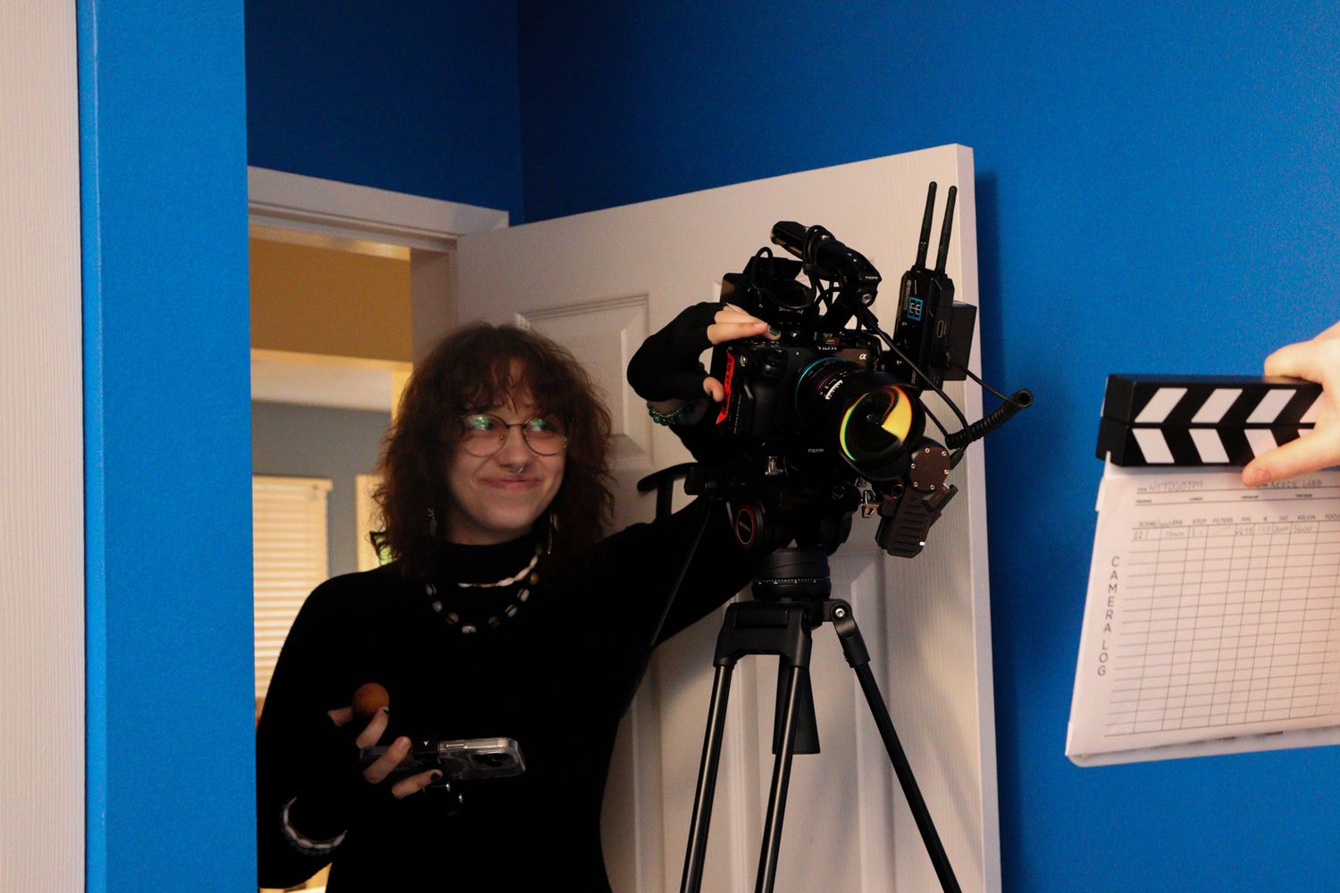 Filmmaker stands beside professional camera on tripod in blue-walled studio with clapperboard visible.