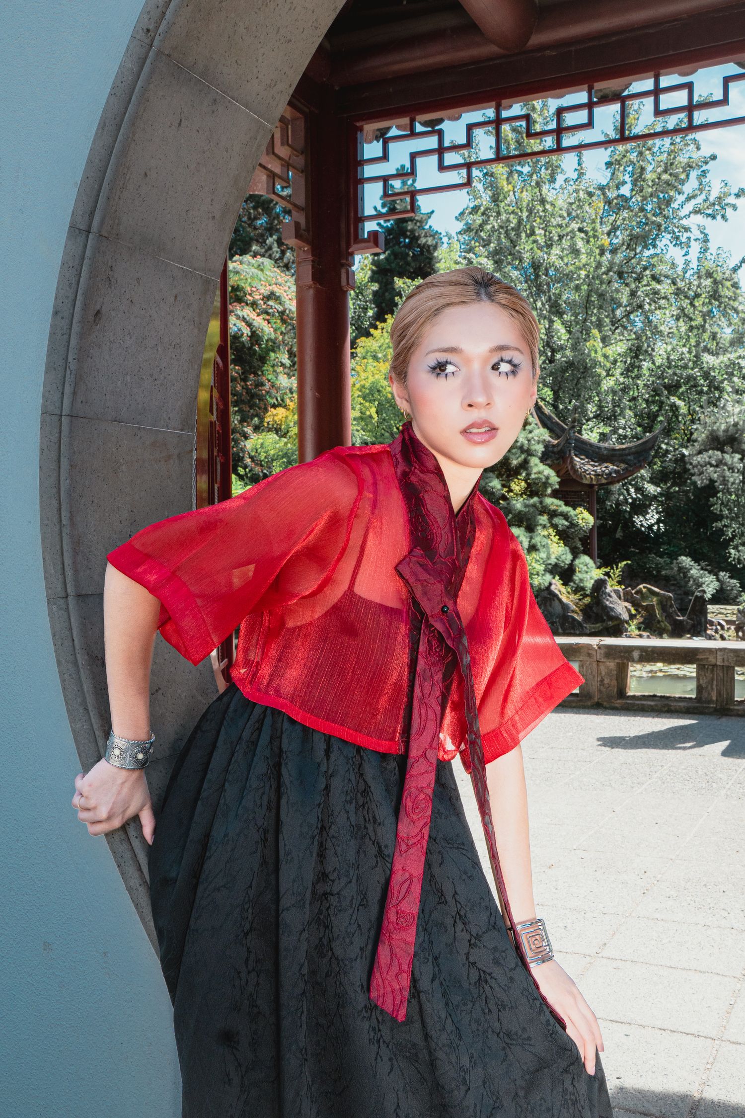 Woman in red sheer top and black skirt posing in a Chinese garden archway with lush greenery.