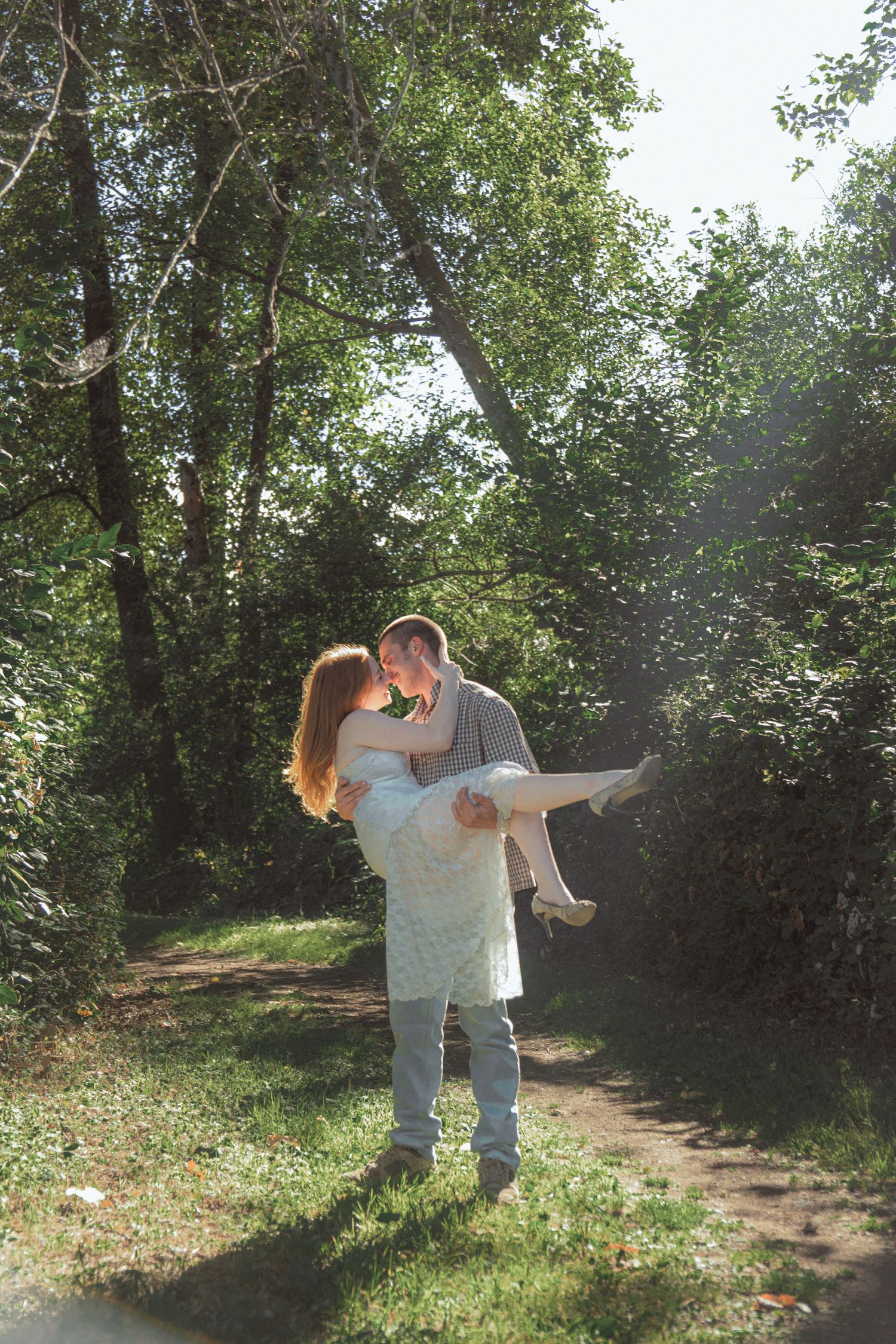 Man lifting woman in his arms for a romantic kiss on a sunny forest path surrounded by lush green trees.
