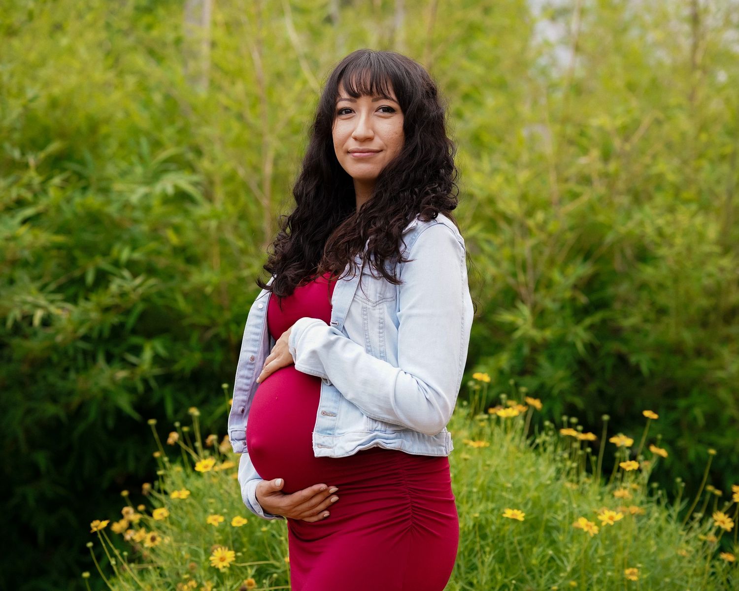 Maternity photoshoot in a garden setting with white jacket and red dress among yellow wildflowers.
