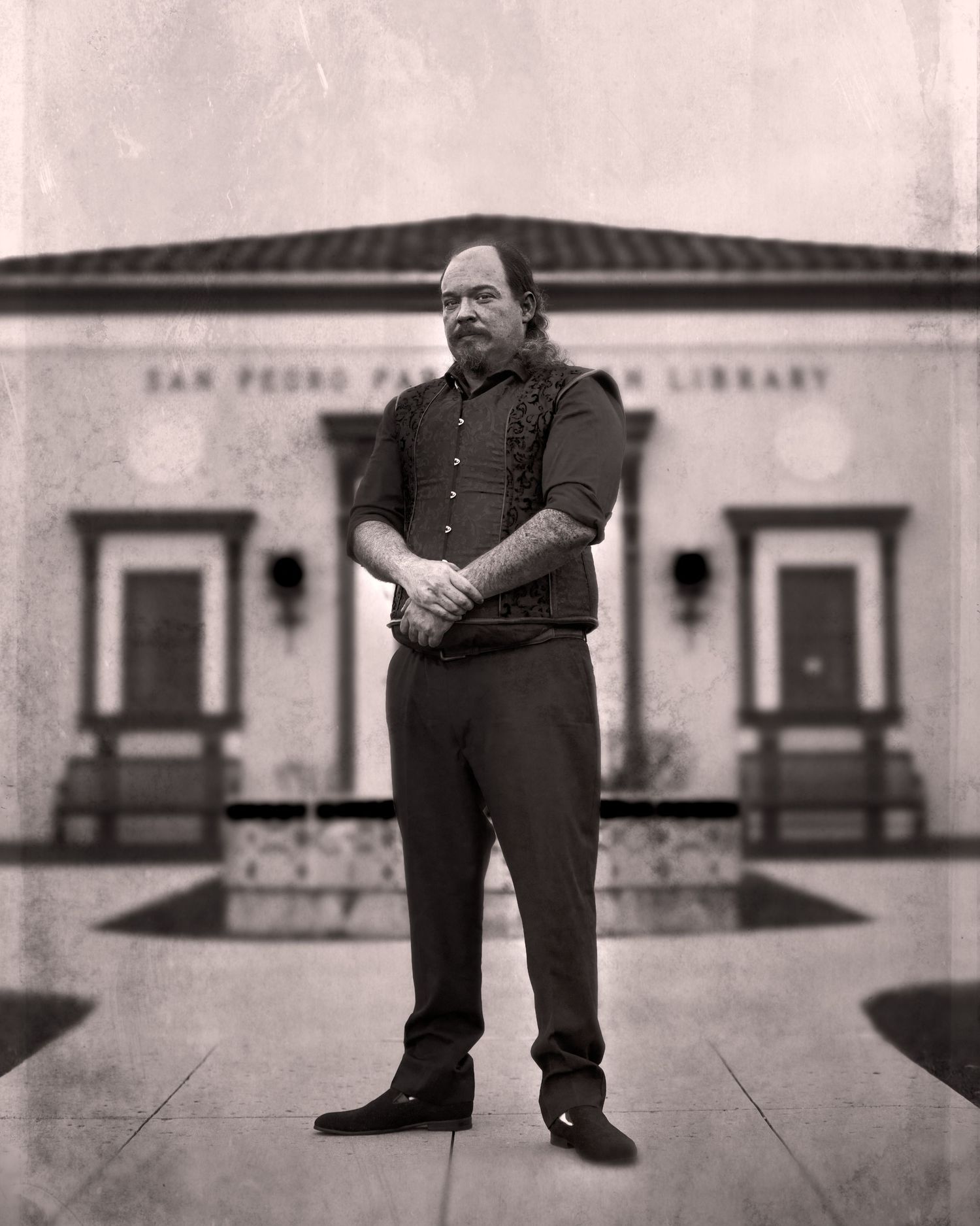Black-and-white street portrait of a man standing in front of a library building with doors and walkway behind him.