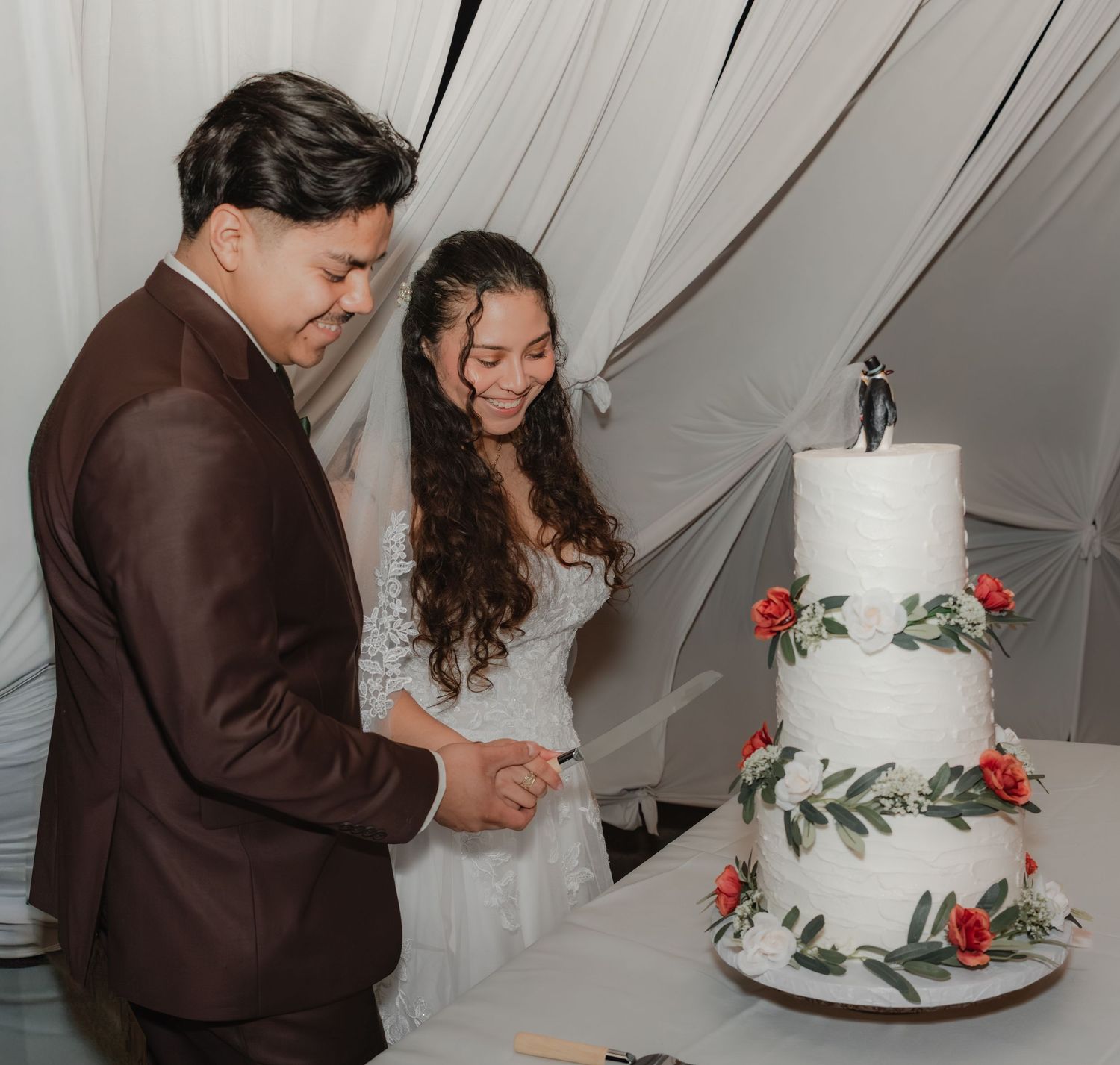 Wedding reception under a white tent—groom and bride smiling while cutting a white floral wedding cake, elegant formalwear, soft warm light