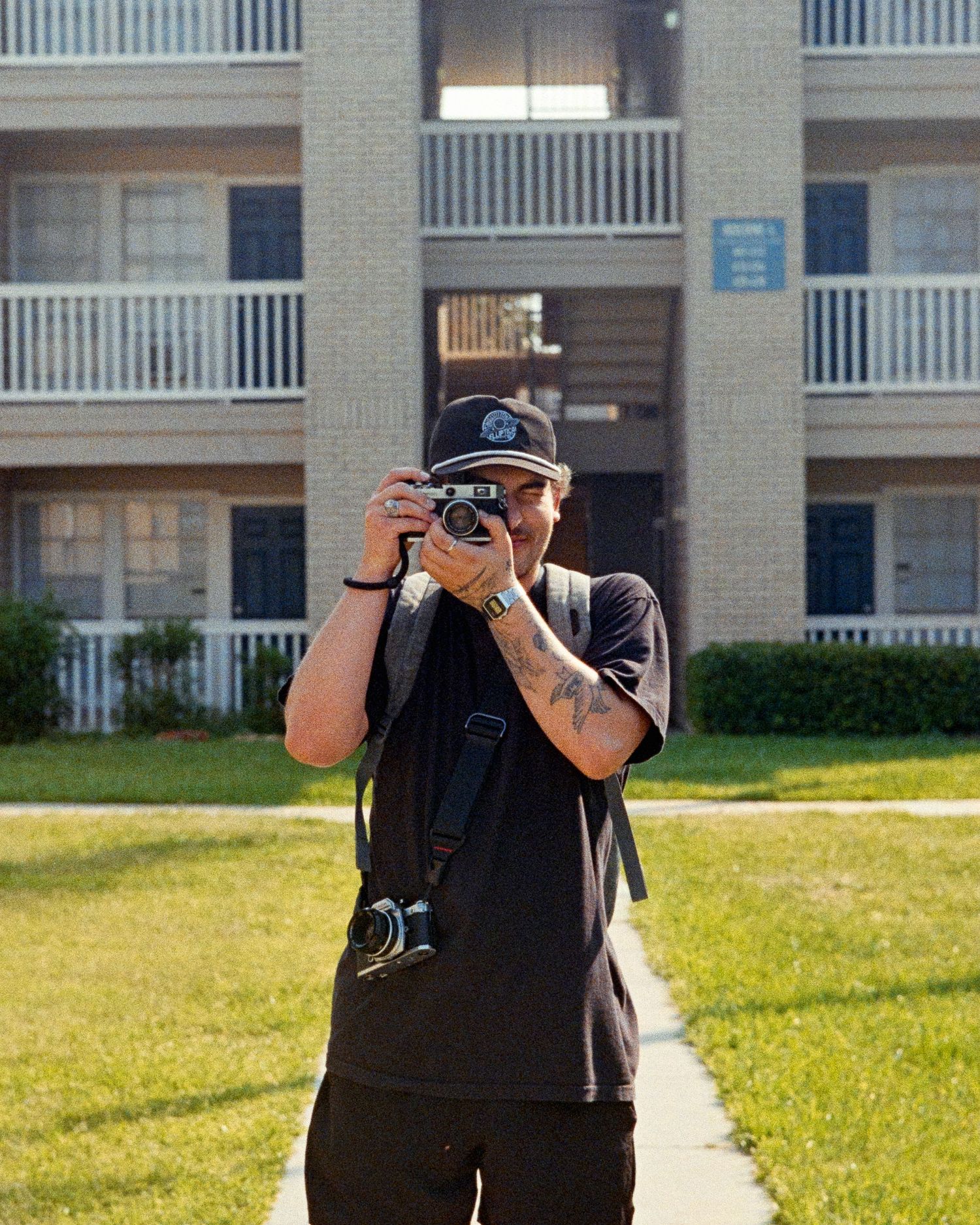 A person in black clothing holding a camera while walking on a path near a brick building with white railings.