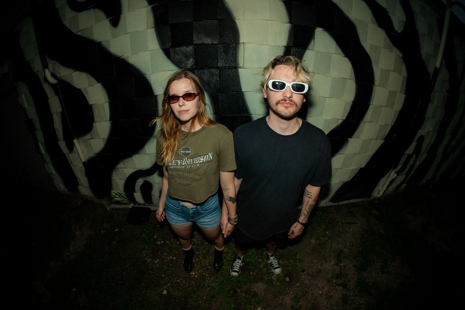 Two people pose against abstract black and white wall pattern in dark lighting.