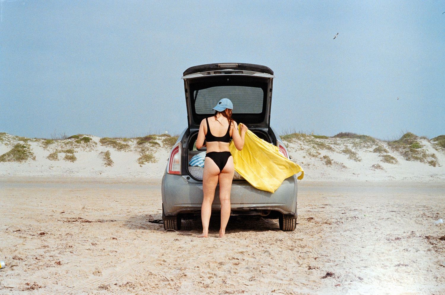 A person in a black swimsuit sits on the open trunk of a car at a sandy beach with a yellow towel nearby.