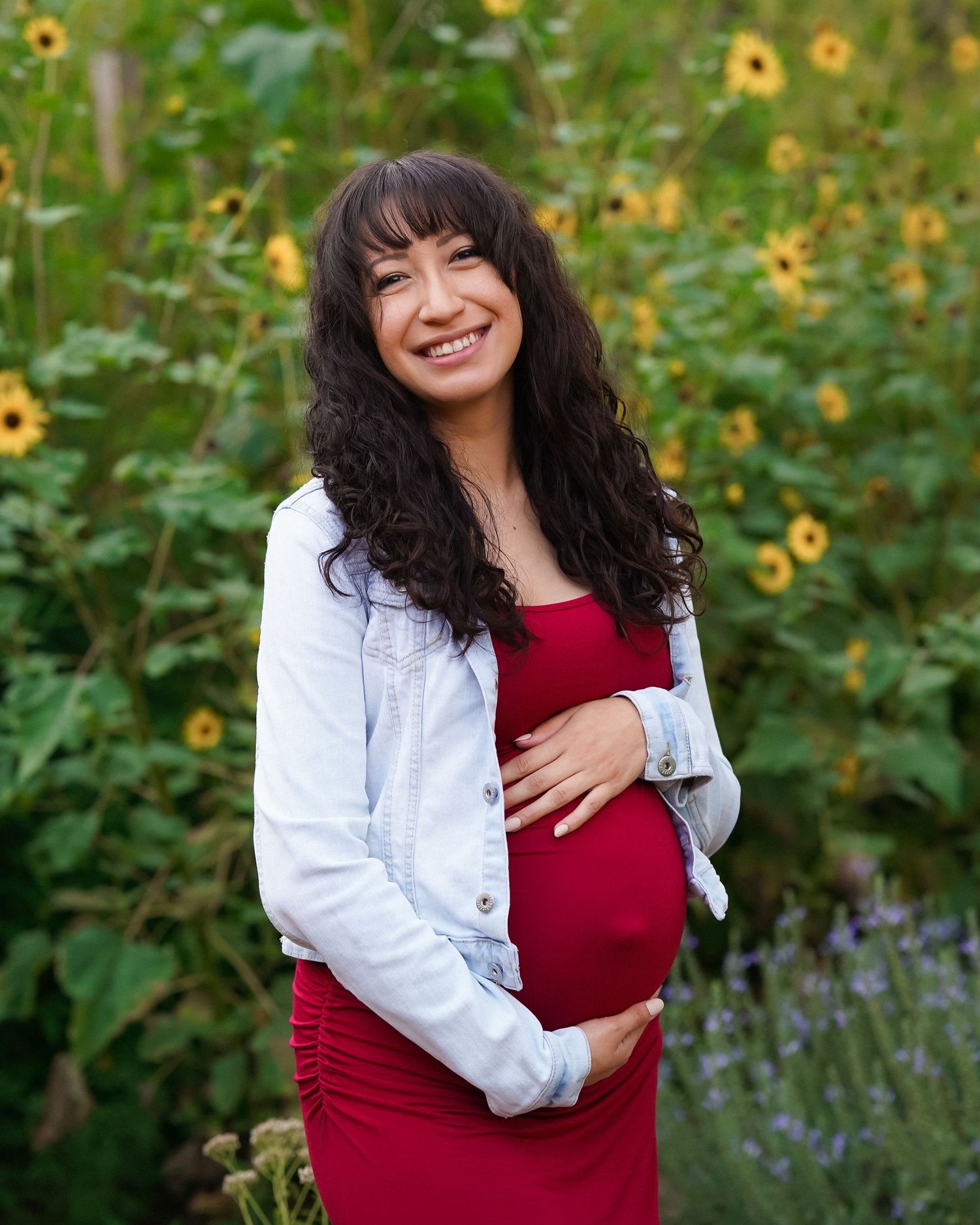 Maternity photos in garden setting with red dress and white jacket among yellow flowers.