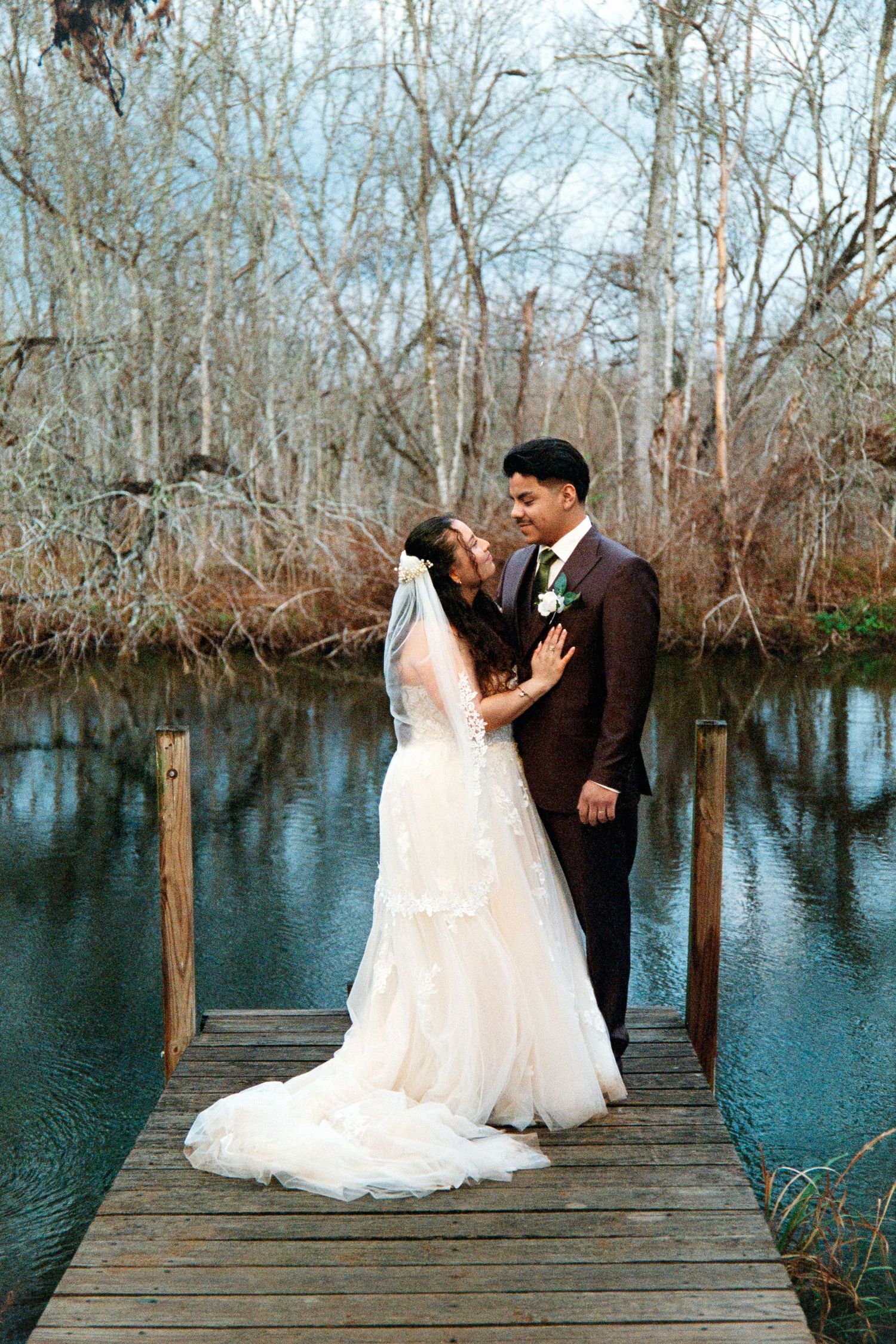A romantic wedding couple shares intimate moments on a wooden dock overlooking a serene lake surrounded by winter trees.