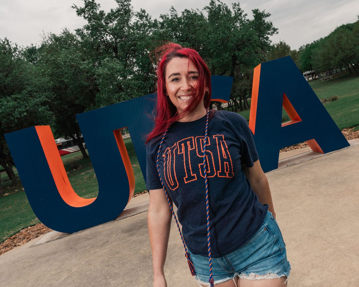 A smiling woman with red hair wearing a UTSA t-shirt stands in front of large UTSA letters on campus.