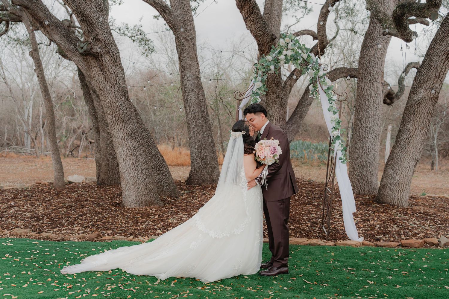 Outdoor garden wedding kiss under a floral arch—bride in a flowing white gown and groom in a dark suit, soft natural light.