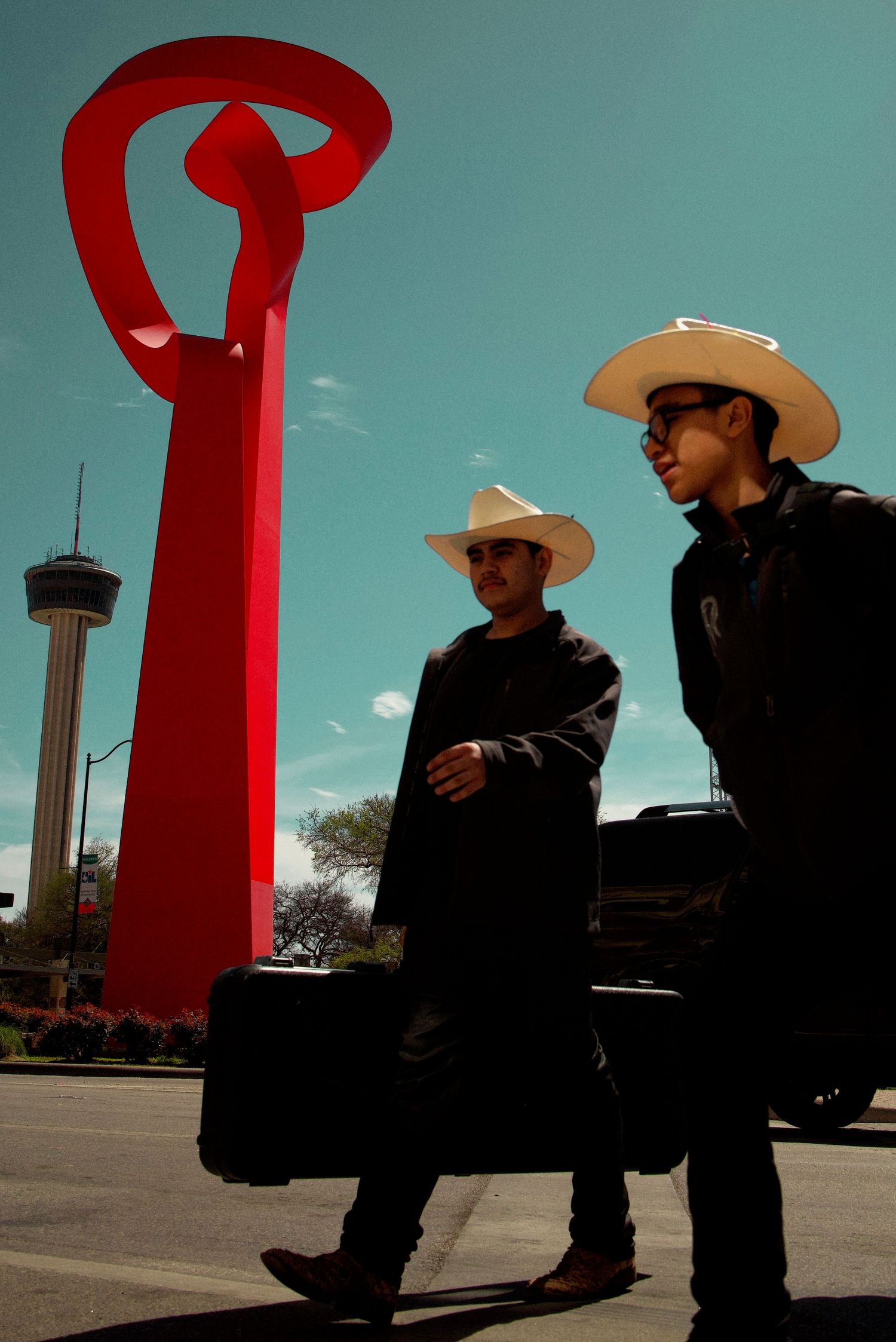 Two people in cowboy hats standing near red sculpture and tower in urban setting.