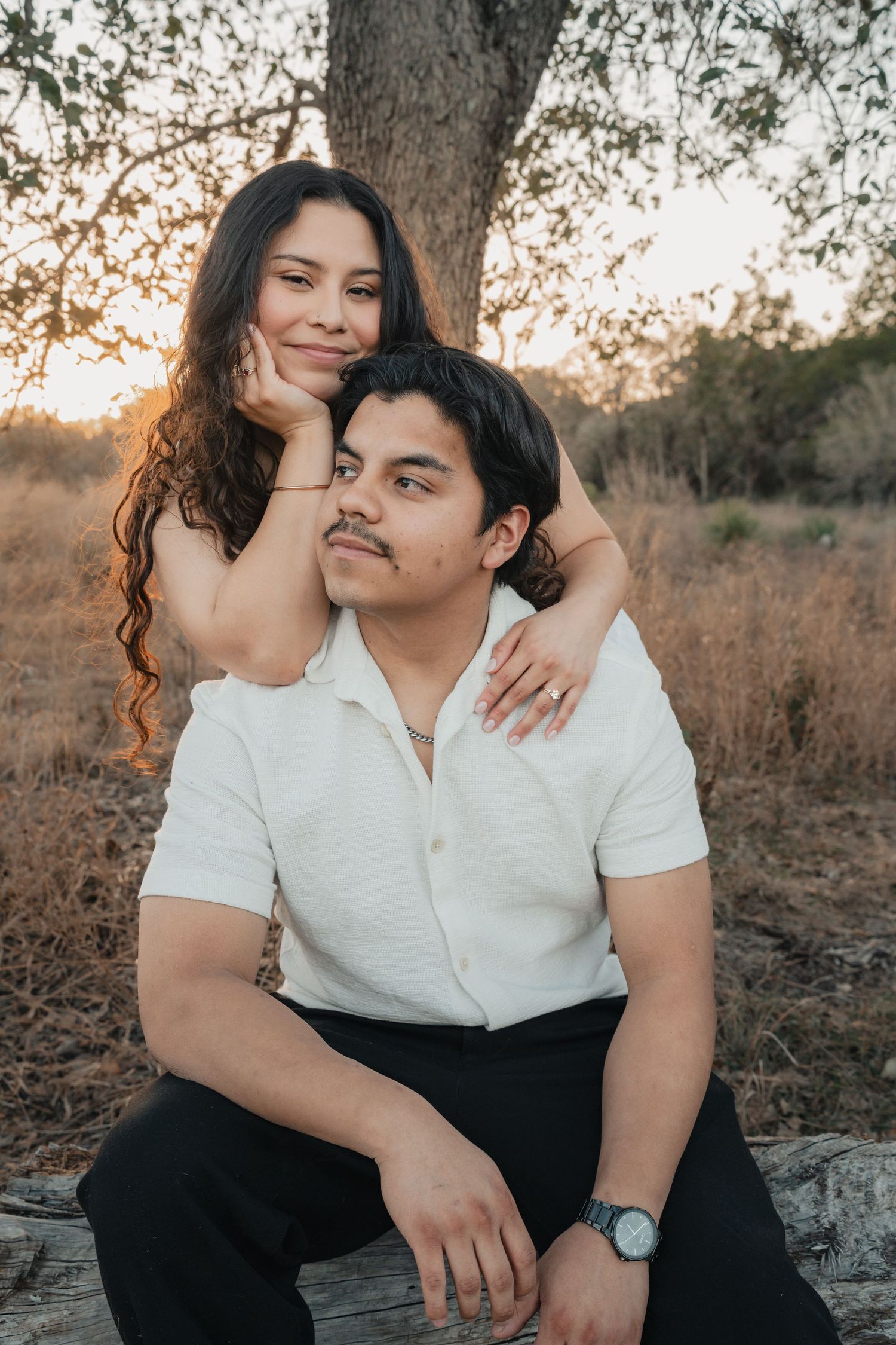 A romantic couple in white shirts embraces while sitting on a fallen tree during golden hour in a rustic outdoor setting.