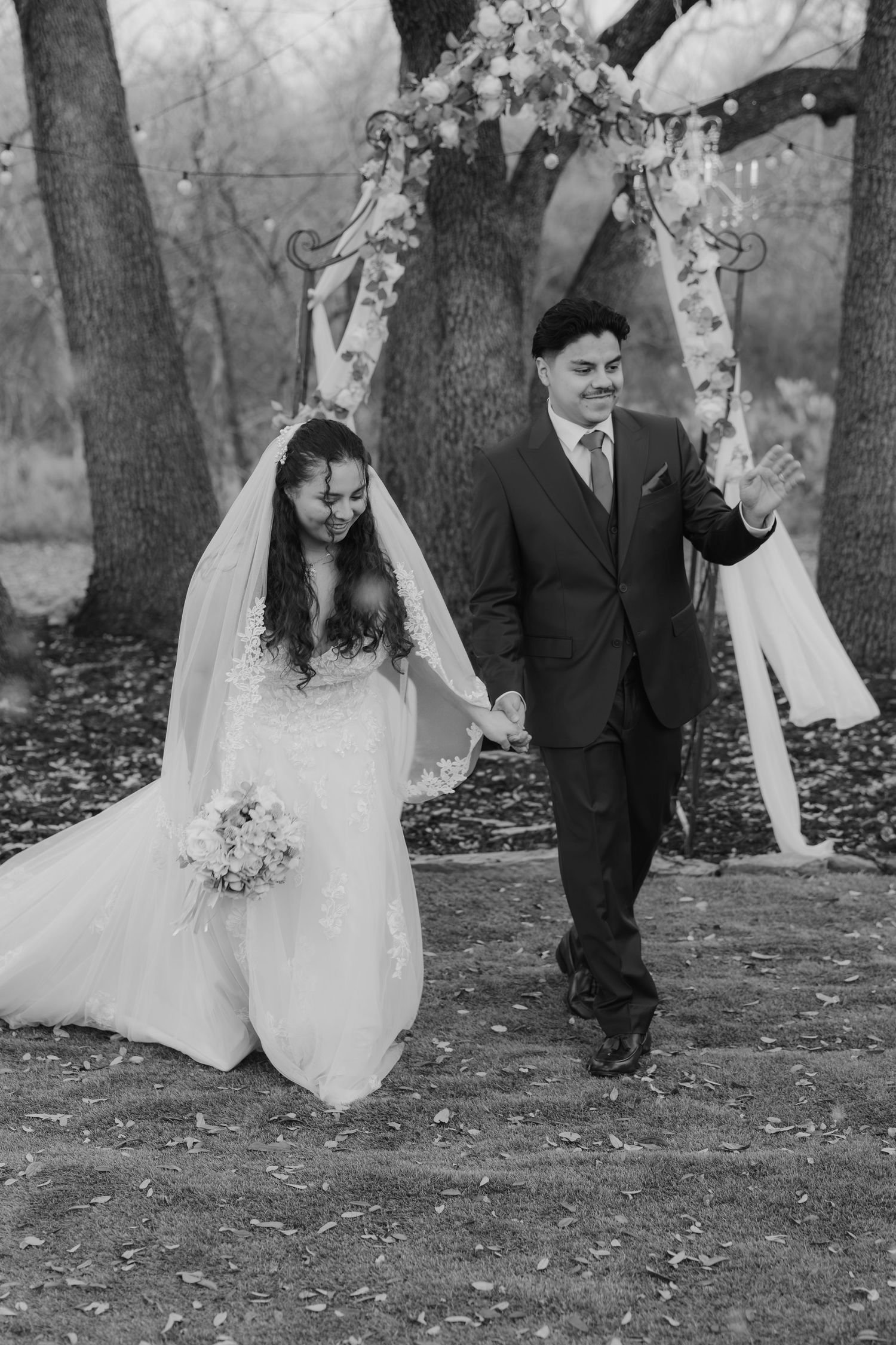 Black-and-white outdoor wedding ceremony—bride in lace gown with veil holding bouquet, groom in suit walking together beneath floral arches.