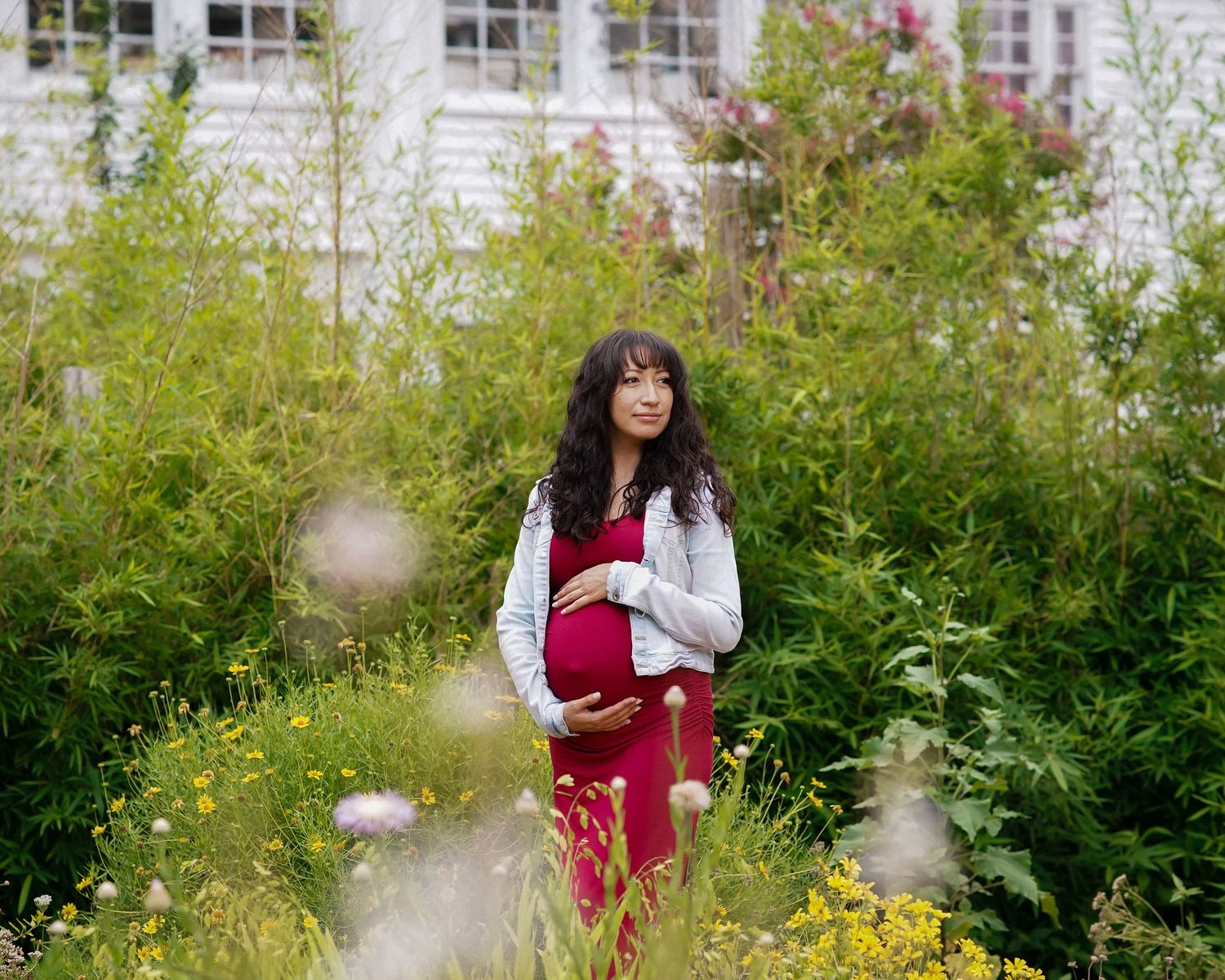 Garden maternity photoshoot with white house and colorful flowers in background.