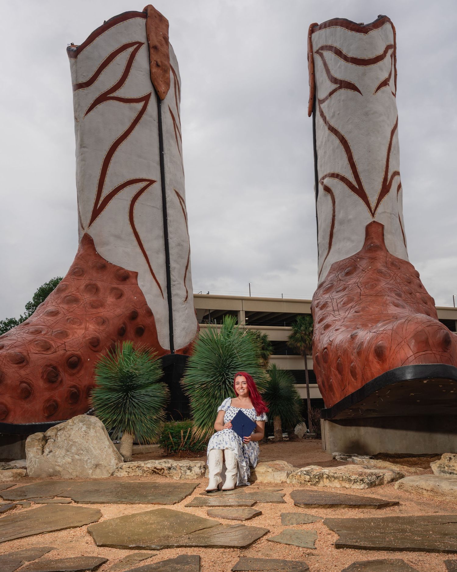Woman with red hair sits between two giant cowboy boots sculpture outdoors on a stone path.