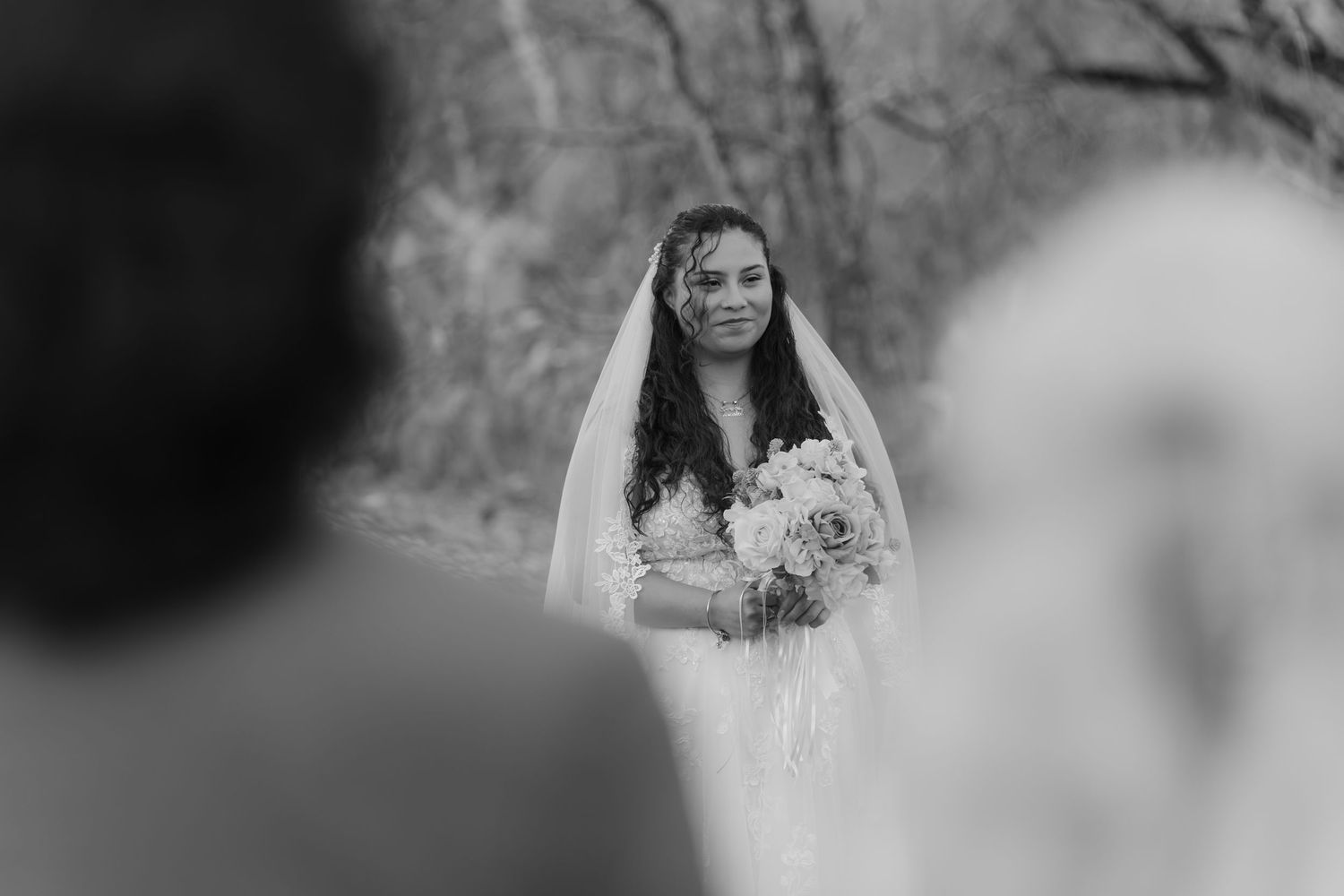 Black-and-white wedding portrait of a bride in lace veil holding a bouquet of white roses outdoors in soft focus.