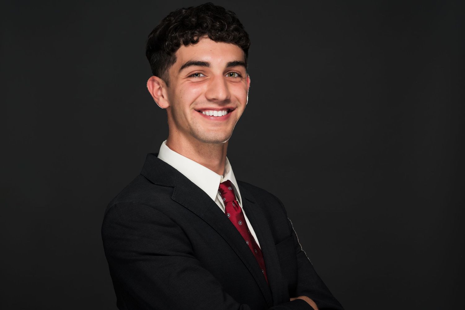 Professional headshot of person in black suit and red tie against dark background.