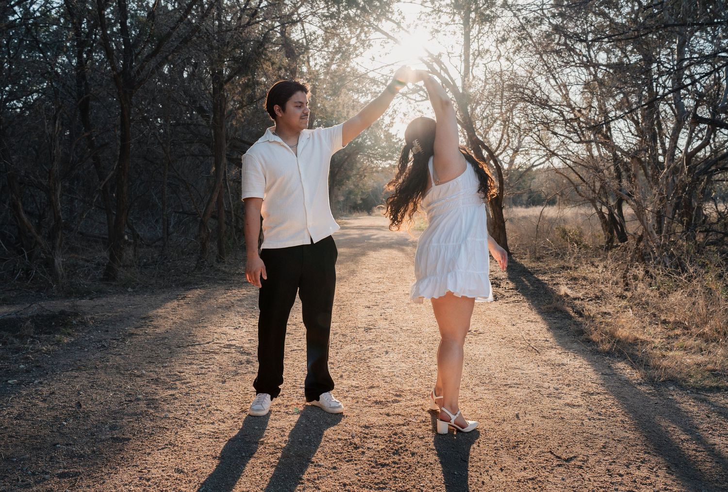 Two people walk together on a sunlit dirt path through trees during golden hour.