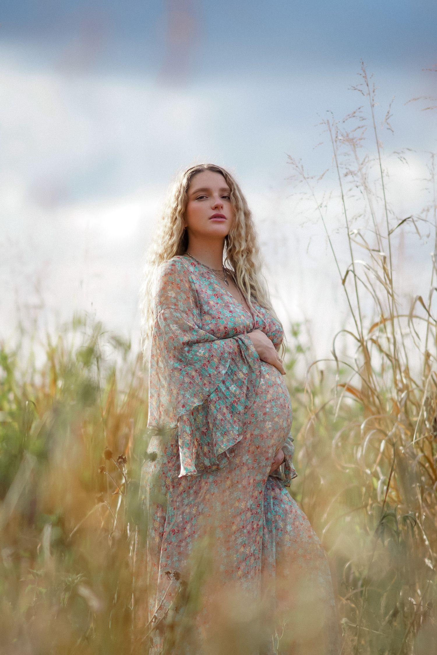 A person in a floral dress poses dreamily among tall grass and wildflowers in soft natural lighting.