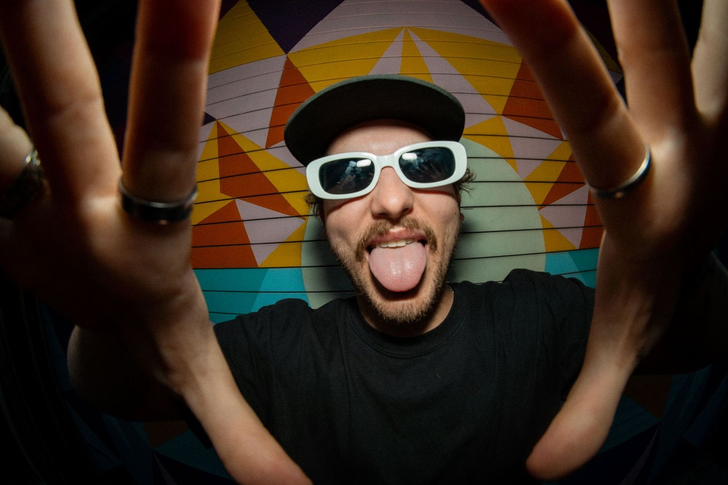 Man in black cap and white sunglasses sticking tongue out with hands spread wide in front of colorful mural.