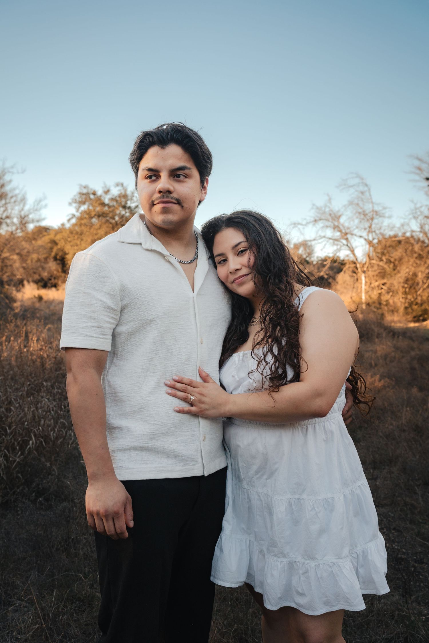A romantic outdoor engagement photo session at sunset shows a couple in white outfits embracing among autumn trees.