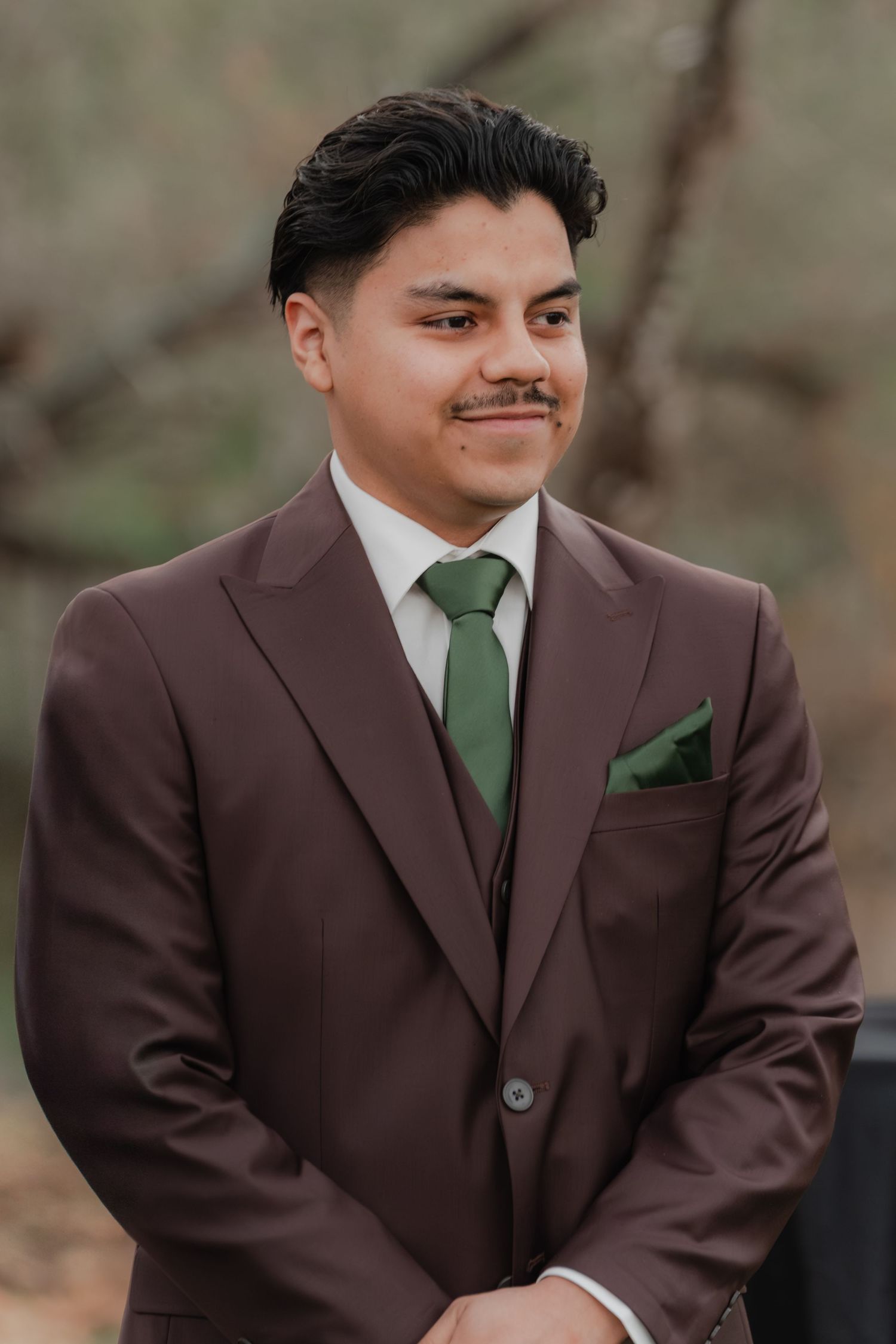 Close-up portrait of a groom in a dark suit with green tie and pocket square, smiling outdoors with soft blurred background.