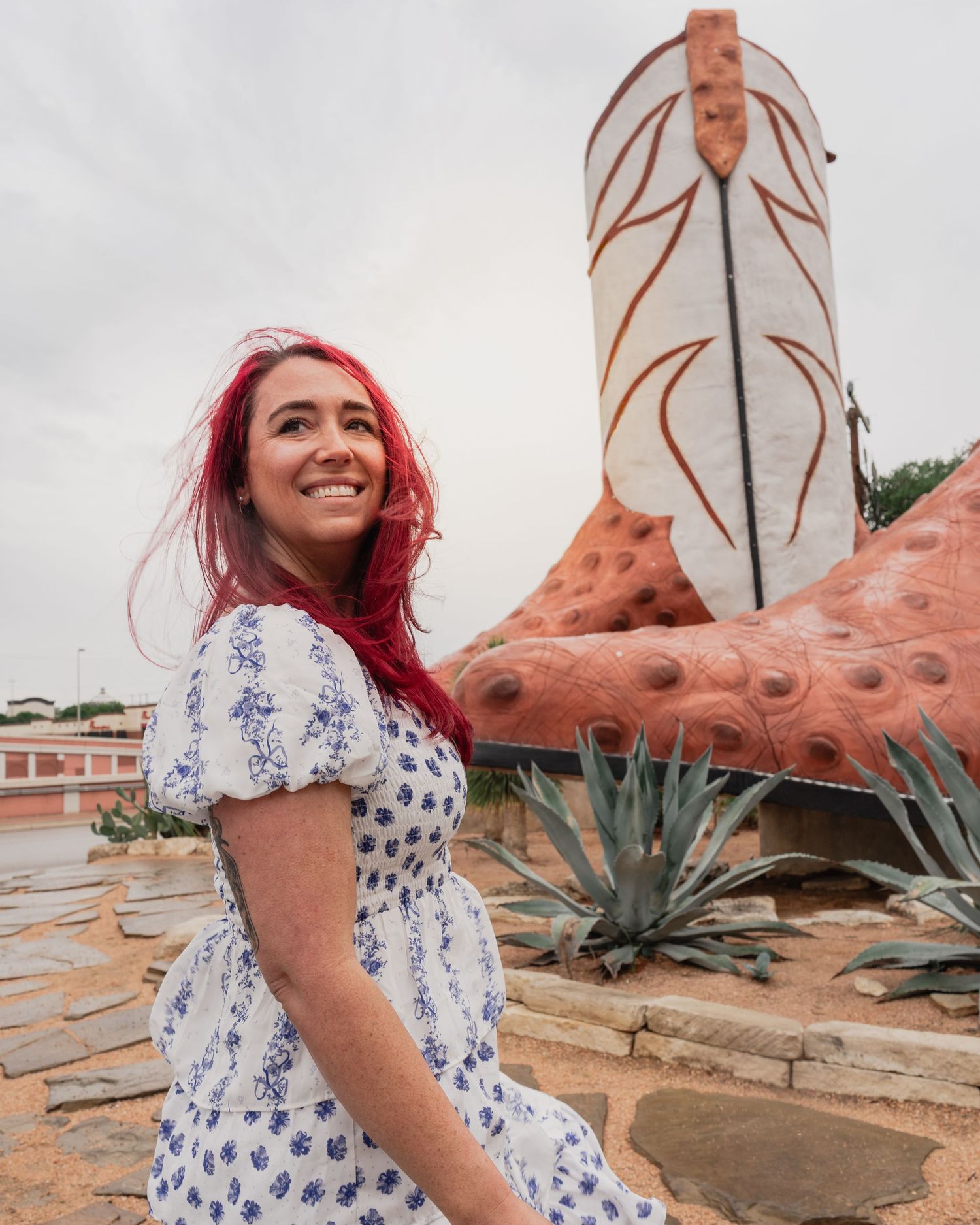 Woman with red hair in floral dress smiling in front of giant cowboy boot sculpture with agave plants.