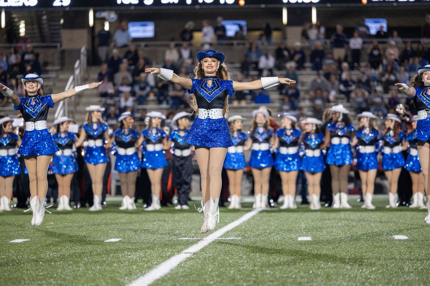 High school dance team members in blue uniforms standing in formation on the field during a night game performance