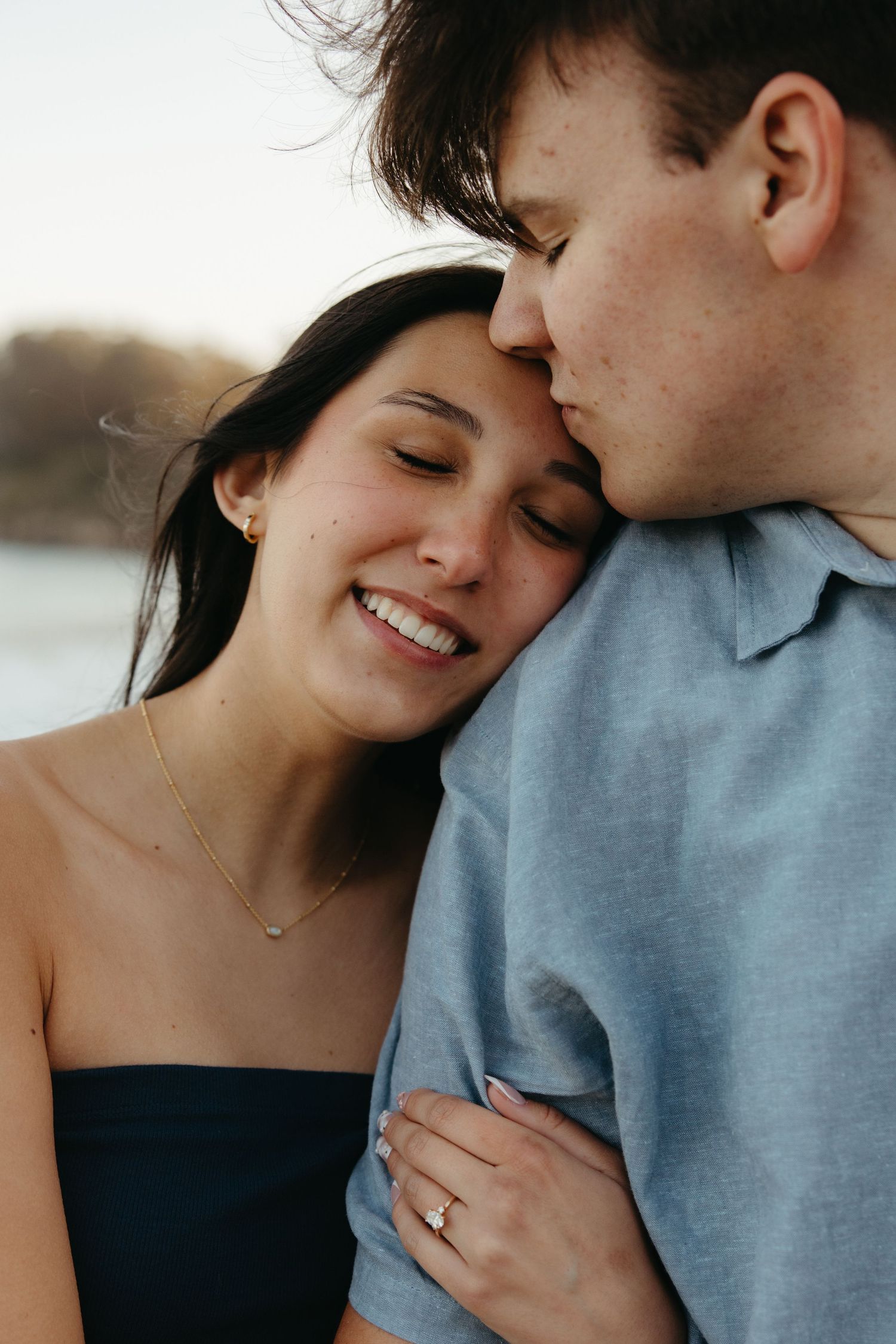 Joyful woman smiling as her partner kisses her temple during a golden hour engagement session.