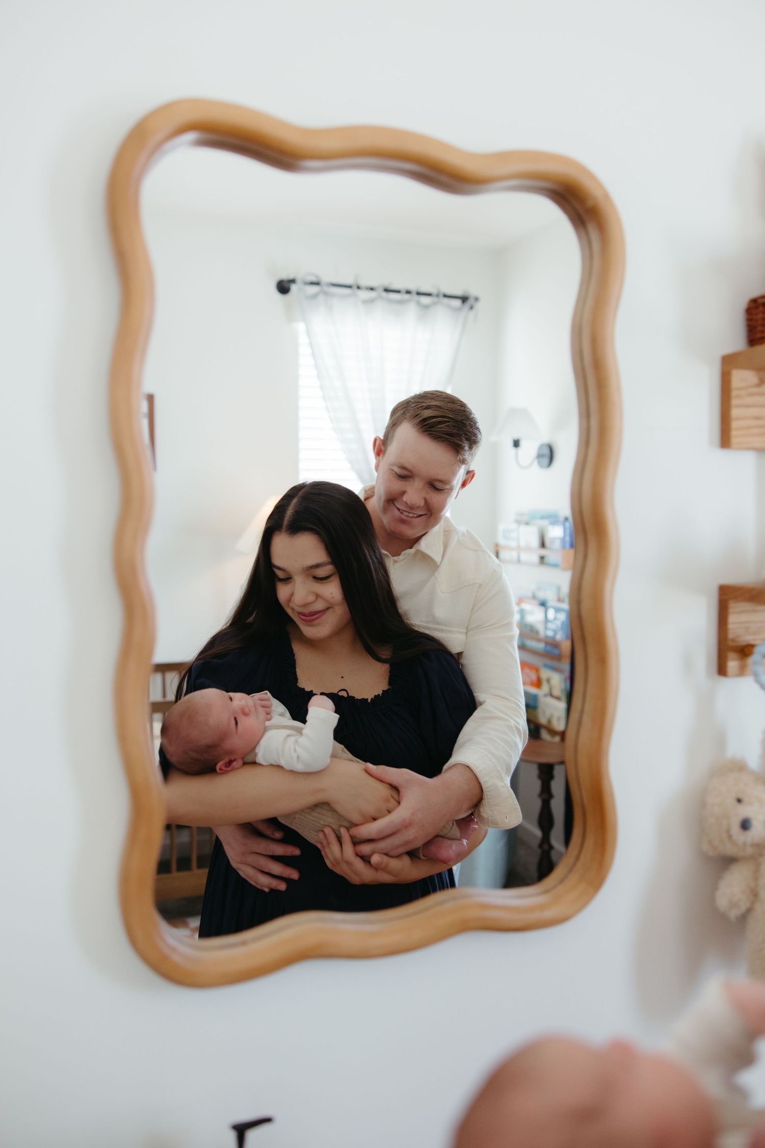 Young couple with newborn baby reflected in a decorative wooden framed mirror on a white wall.