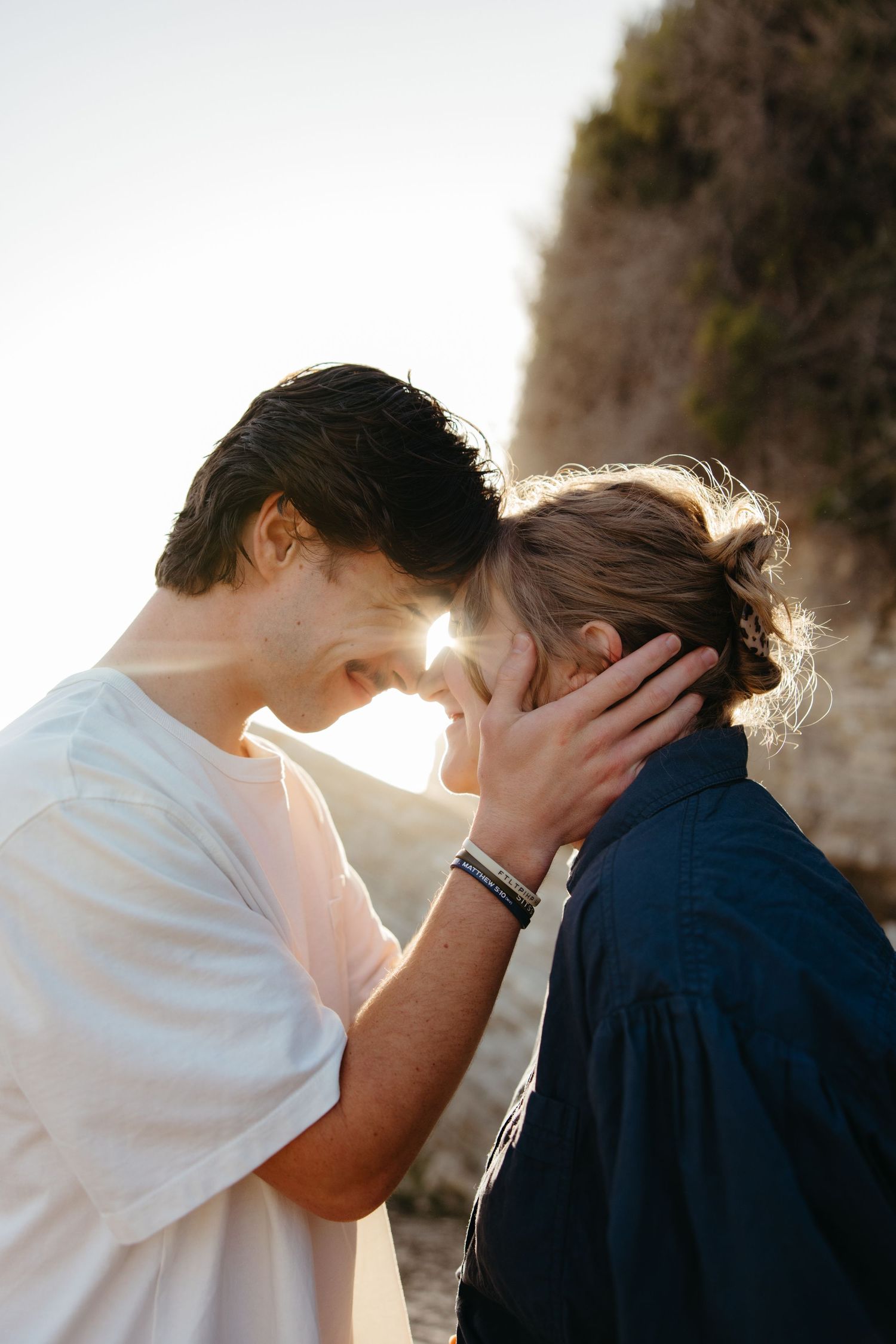 Couple tenderly touch foreheads with sun flare behind them near rocky coastal cliffs.